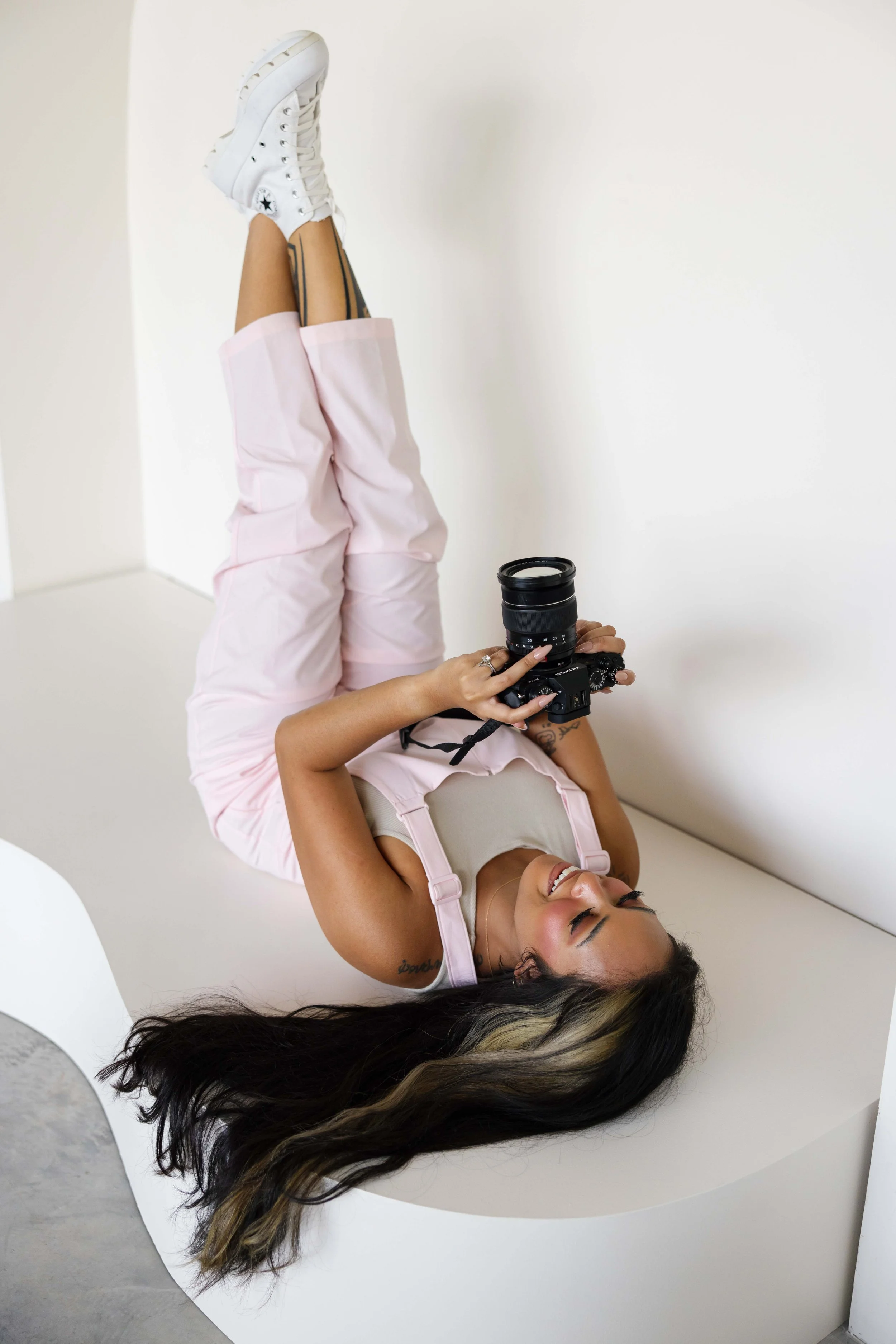 A smiling woman with tattoos lying on her back on a white curved surface, holding a camera, with her legs raised and crossed at the ankles, wearing white sneakers, pink pants, and a beige top.