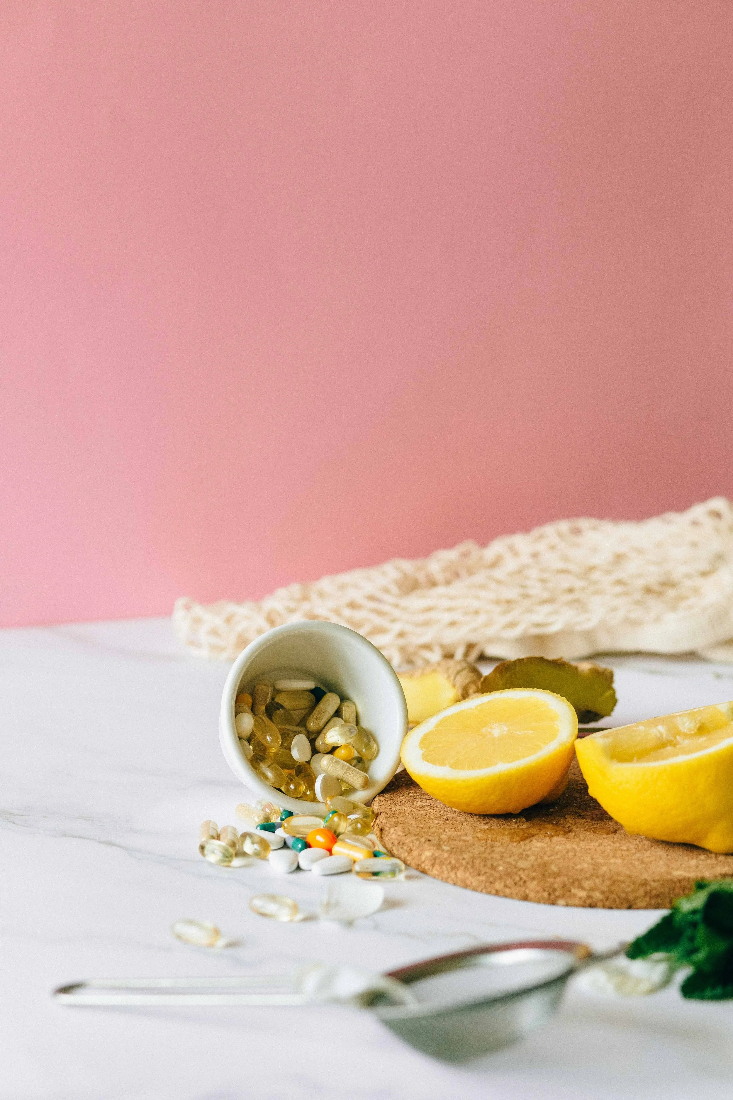 A small cup of assorted pills and capsules spilled on a white surface next to lemon wedges, a wooden cutting board, a crochet cloth, and some herbs, set against a pink background.
