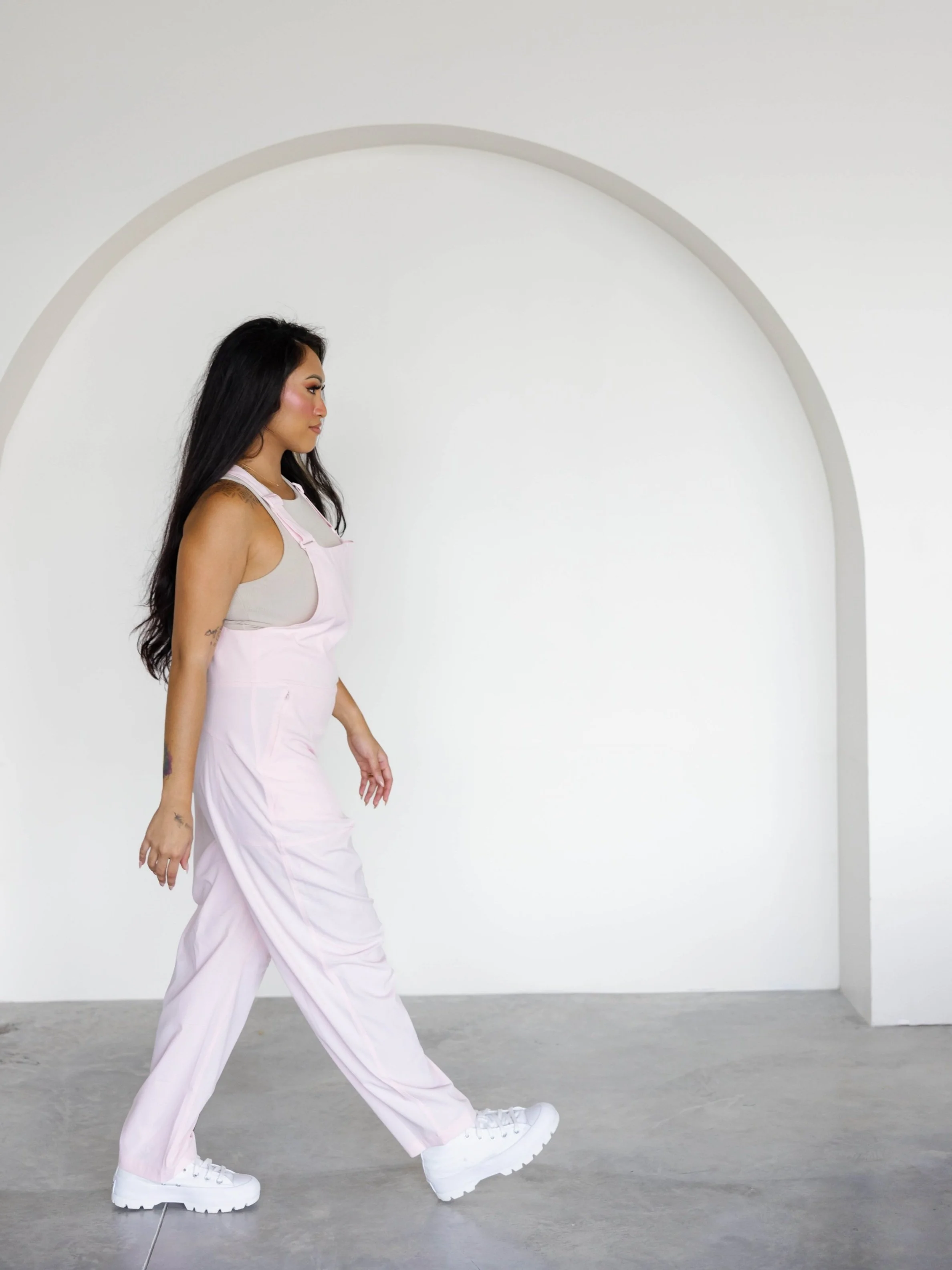 A woman with long dark hair walking past a white wall with a large arch opening.