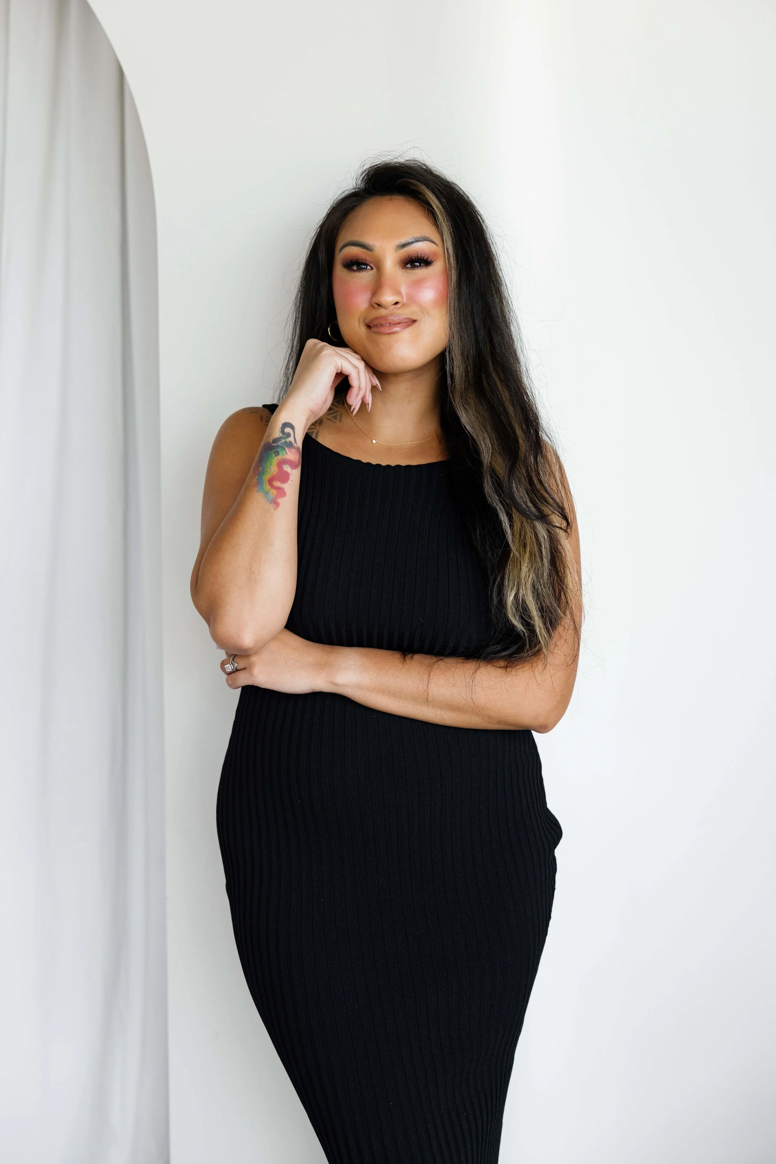 A woman in a sleeveless black ribbed dress posing with her right hand near her chin against a plain white background.