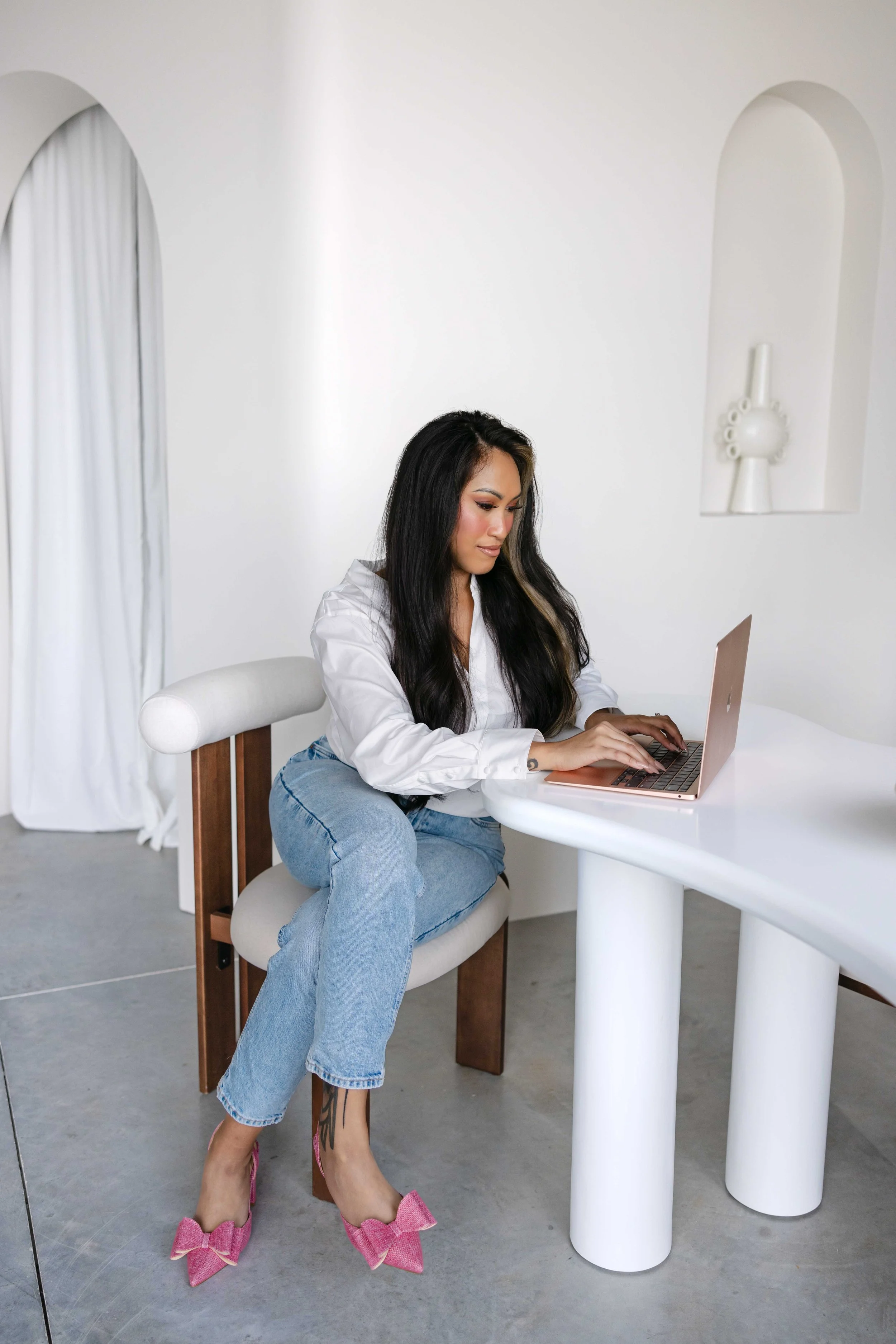 Woman sitting at a white table working on a pink laptop in a minimalist, white room with decorative wall art.