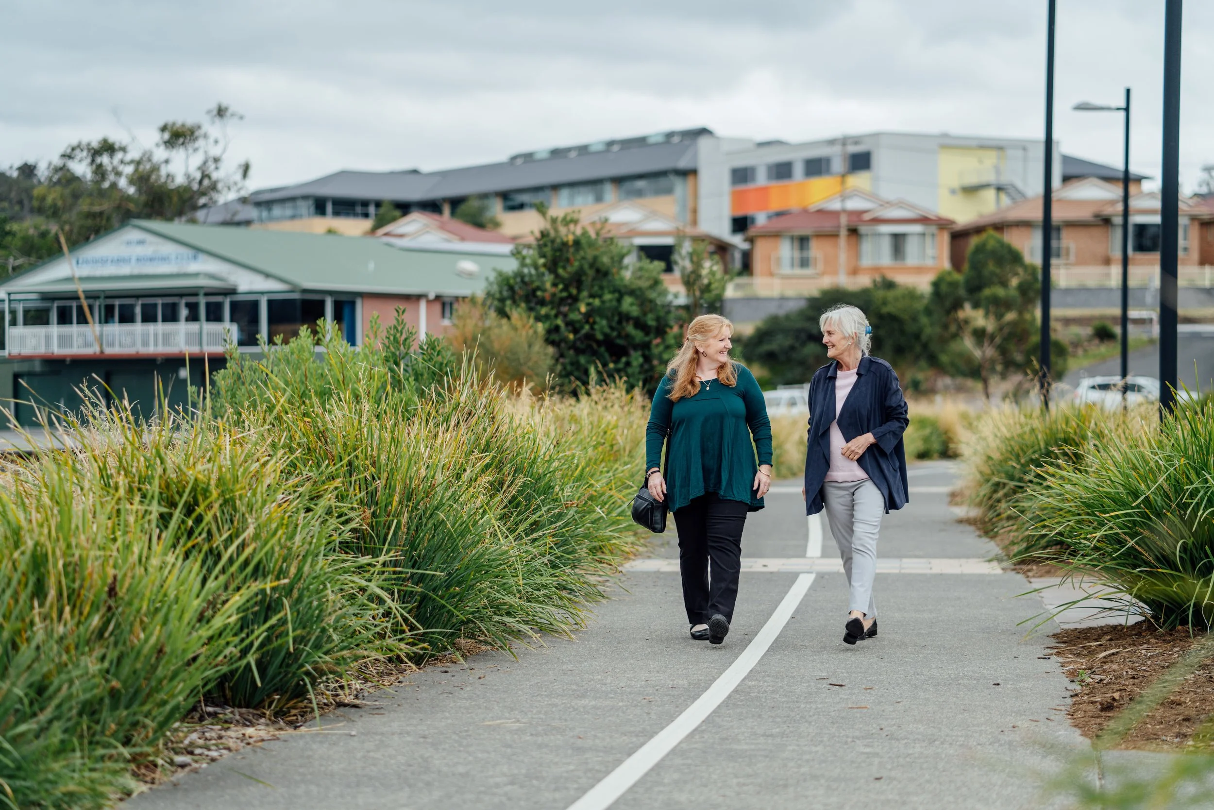 Two women walking and talking on a paved pathway lined with tall green plants, with houses and buildings in the background.