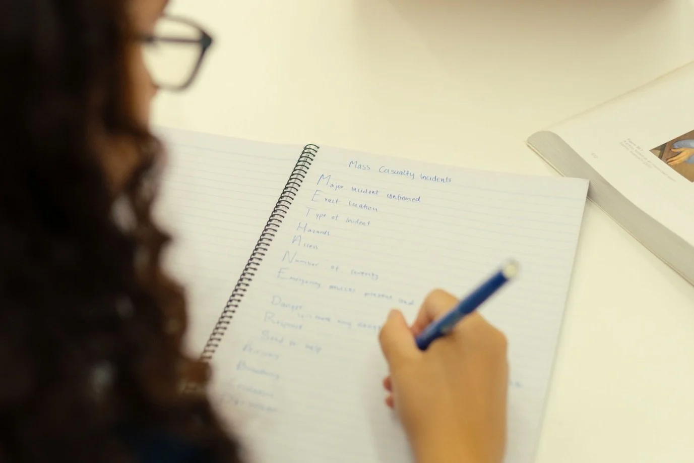 A person with curly hair and glasses writing notes in a notebook with a blue pen.