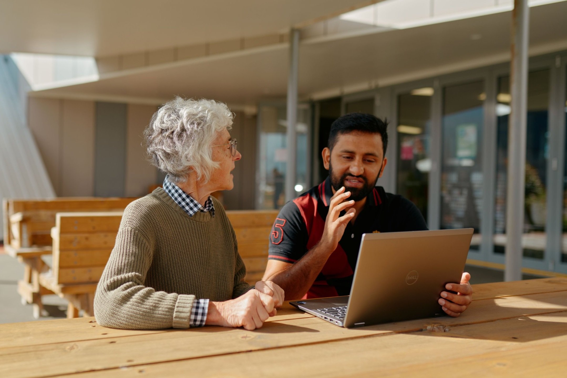 A woman and a man sitting at a wooden table outdoors, looking at a laptop together, engaged in conversation.