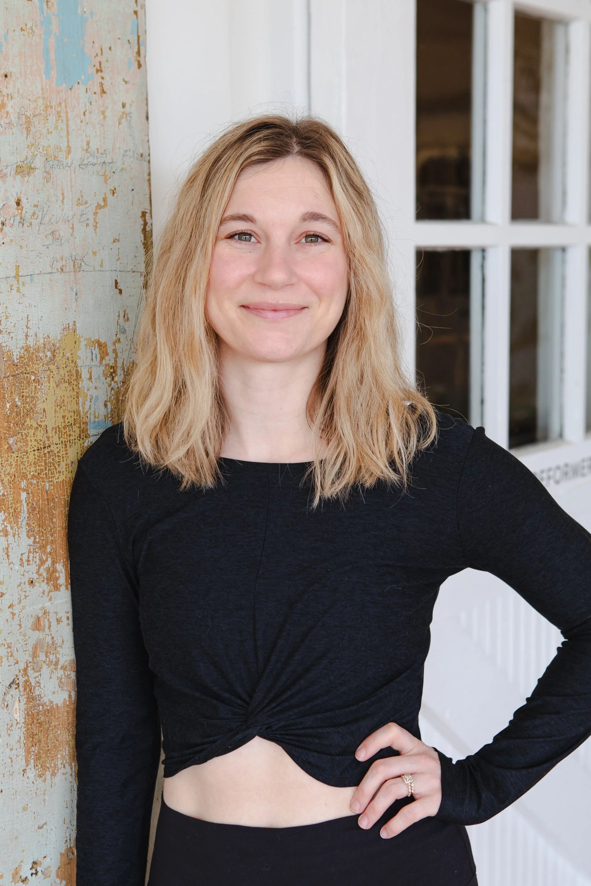 A woman with blonde hair sitting cross-legged on a wooden bench with a beige pillow behind her, looking at the camera and smiling.