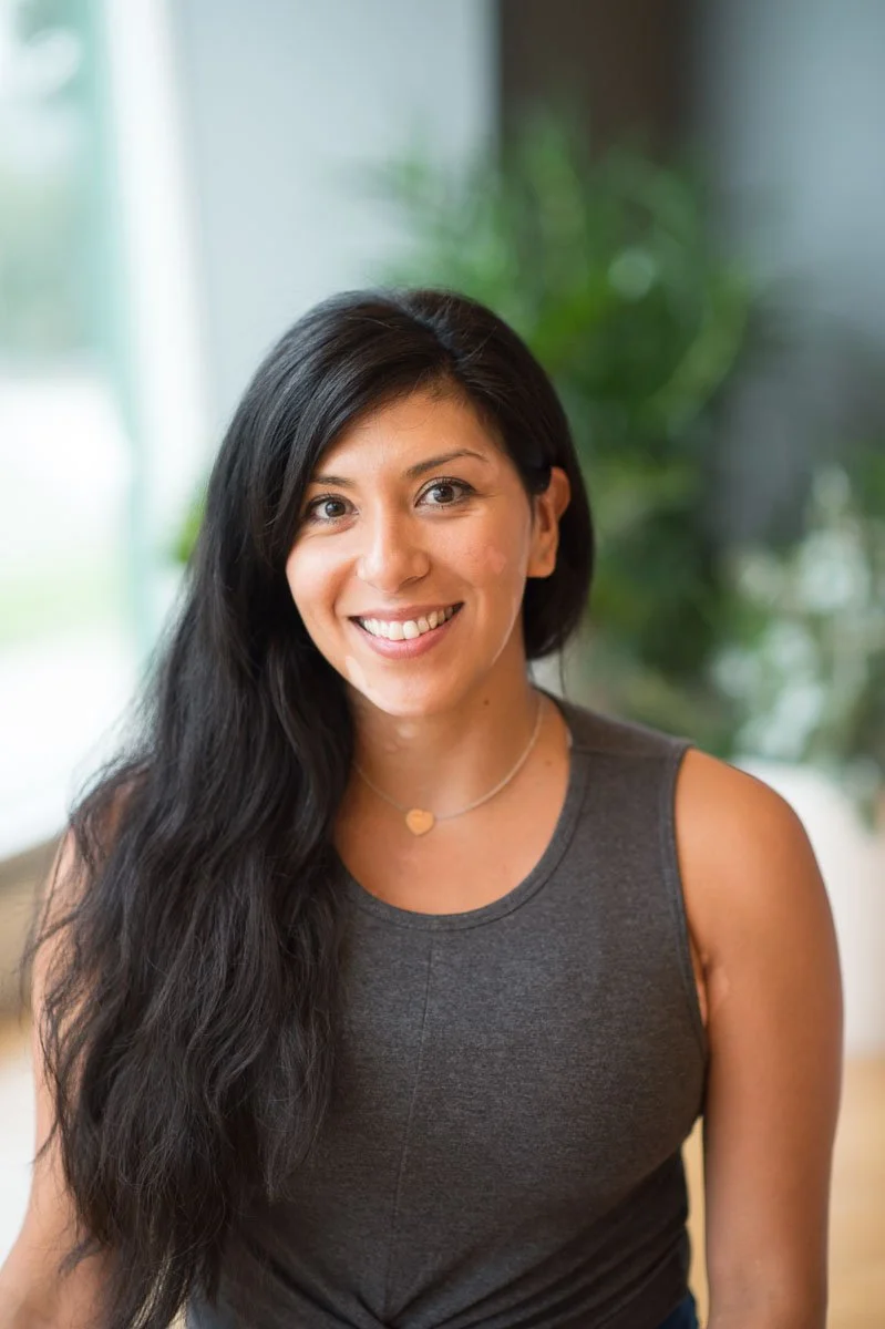 A woman with long dark hair, wearing a blue sleeveless top and gray pants, sitting on a bench in a room with wooden flooring and brick wall background, smiling at the camera.