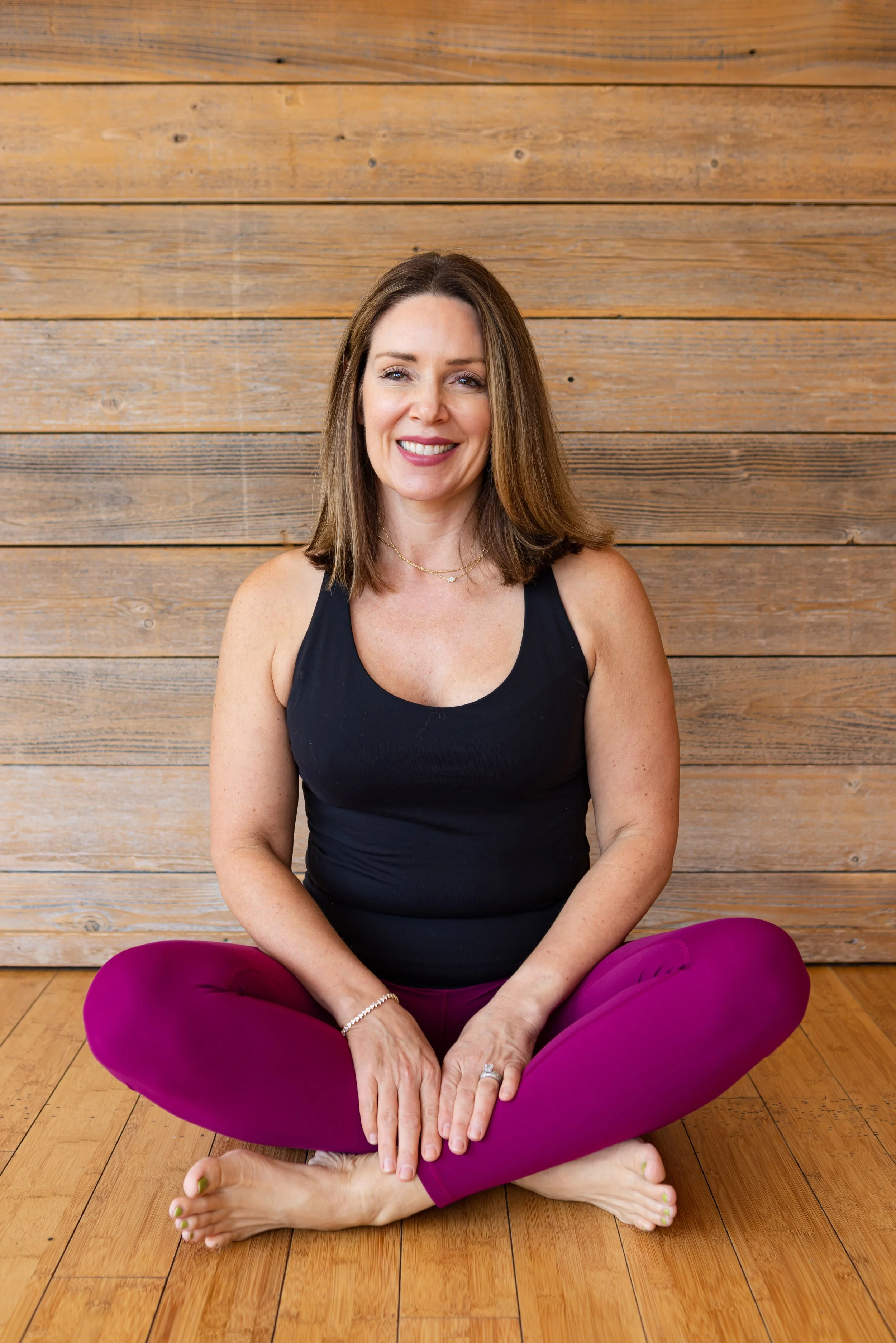 A woman with long dark hair and a bright smile, wearing a black top, indoors with a brick wall and wooden floors.