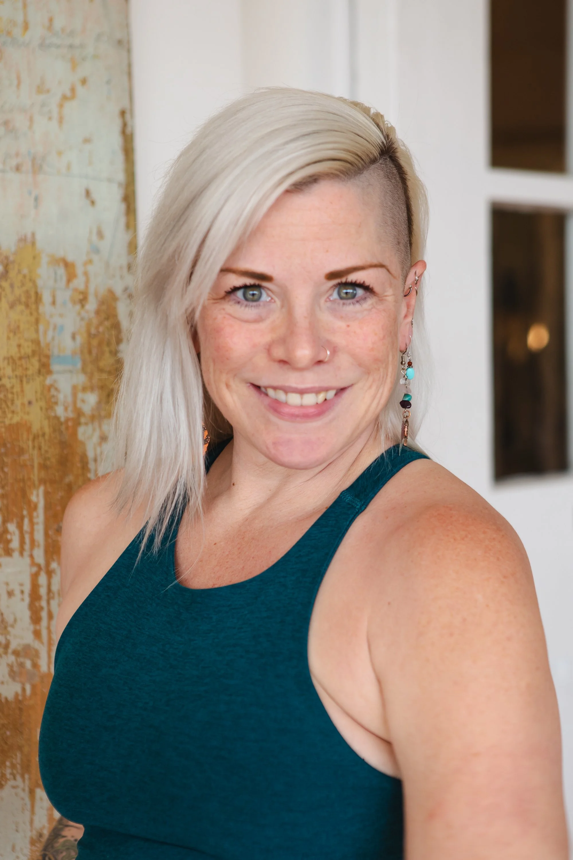 A woman with short gray hair stretching her arms overhead in a fitness studio, wearing a red sports top and black shorts, standing in front of a light green wall and wooden rail, with a potted green plant on the floor.