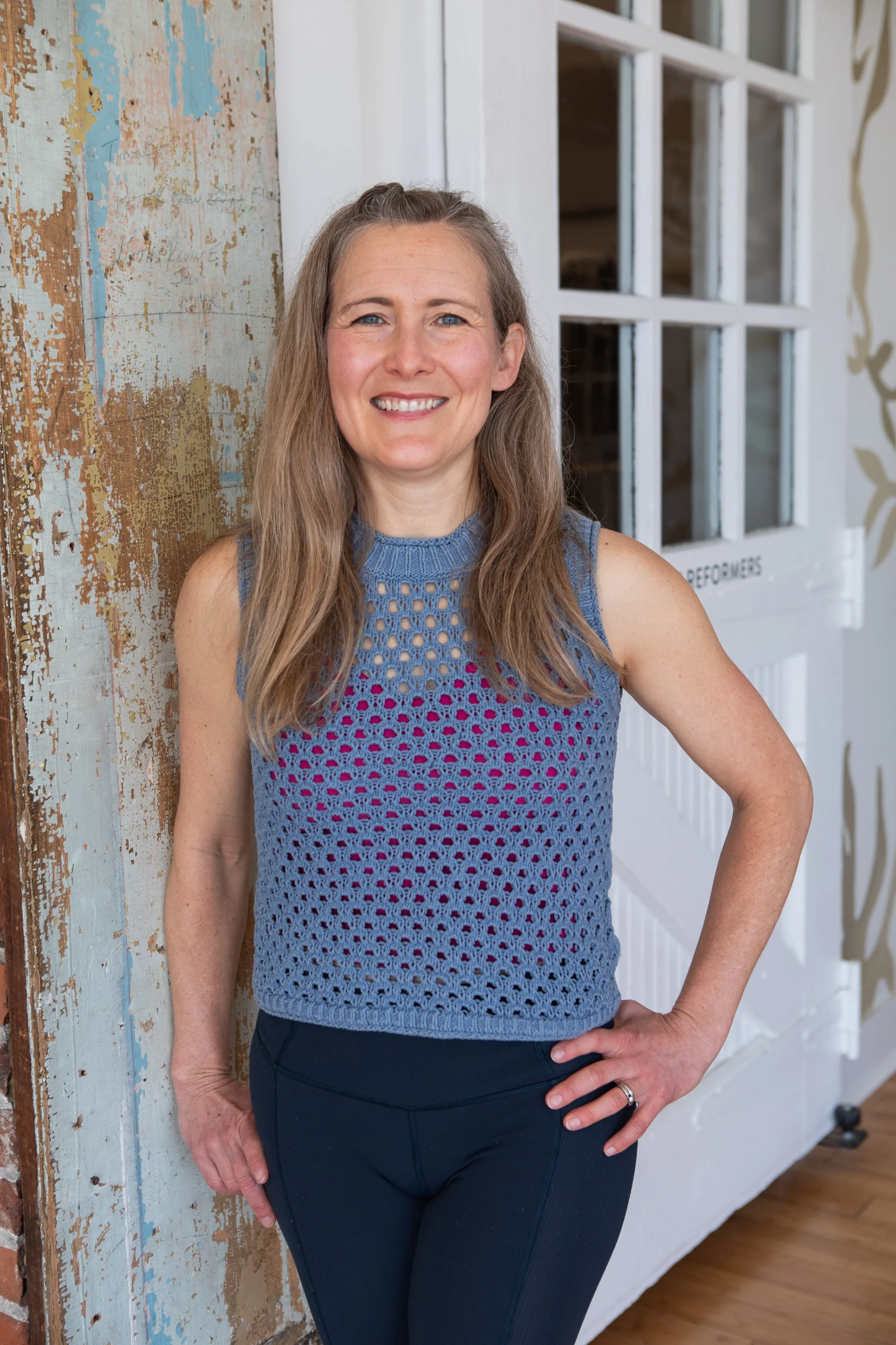 A woman wearing a light green tank top and black pants, smiling and standing indoors next to a wall with exposed brick and insulation.
