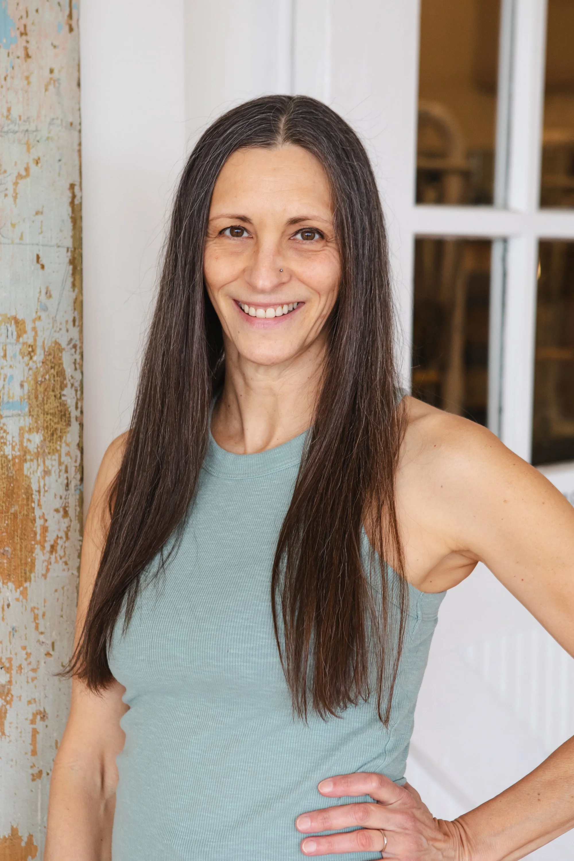 A smiling woman with long dark hair wearing a blue sleeveless top and gray leggings, sitting on a striped cushioned seat in a room with wooden floors and a brick wall in the background.