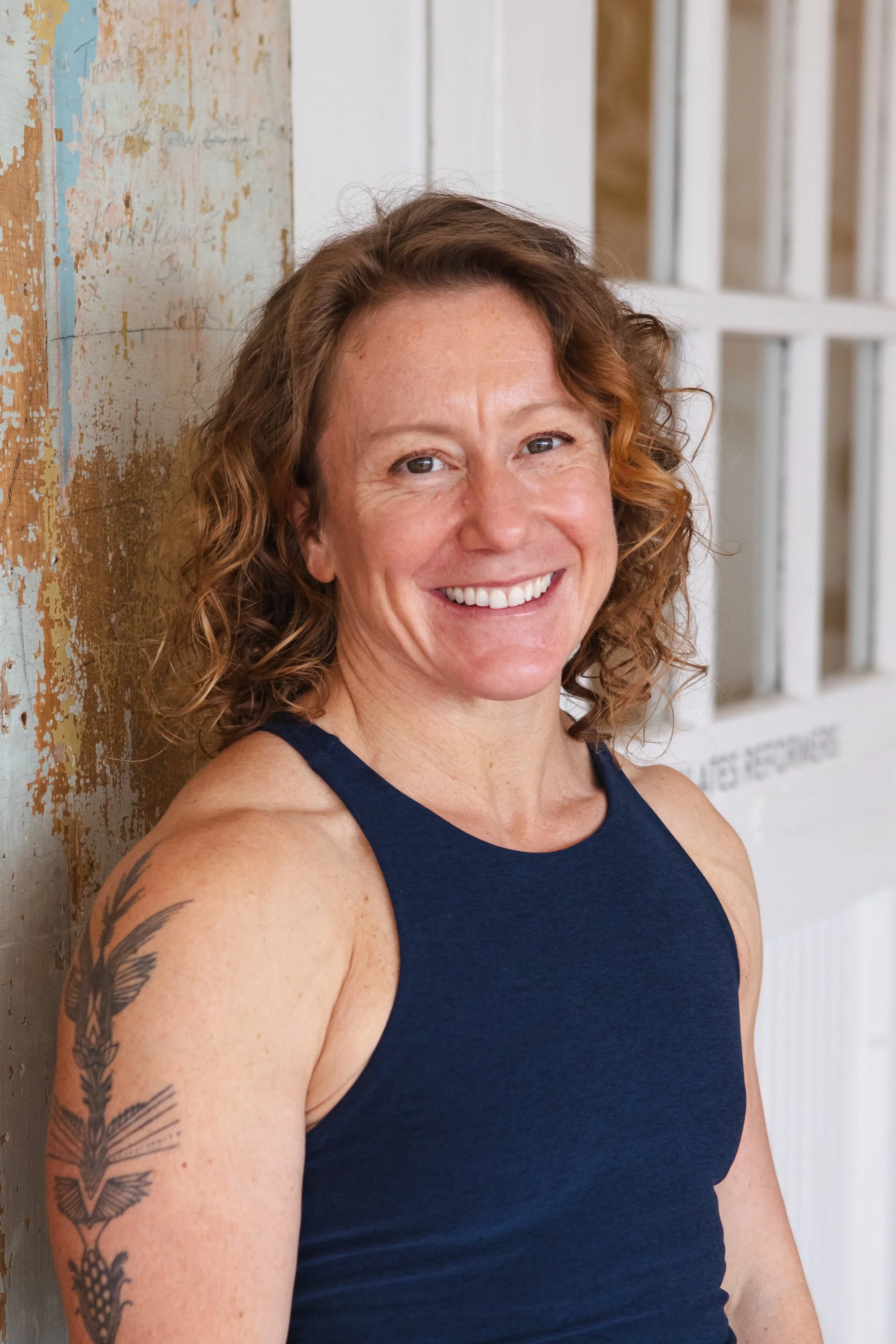 A red-haired woman smiling, wearing a blue athletic tank top and black workout leggings, standing outdoors beside a brick wall and a white door.