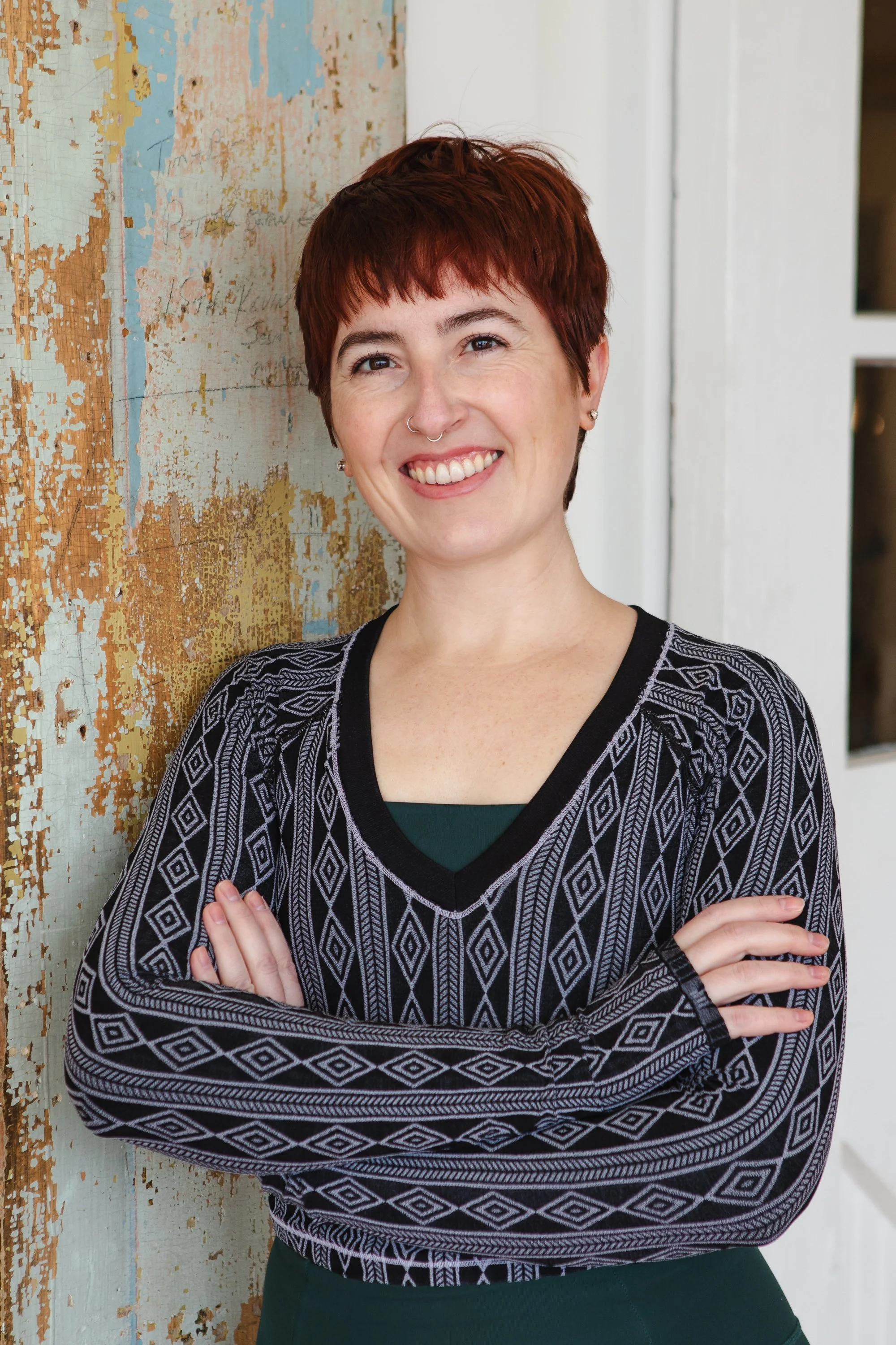 Smiling woman with short brown hair and a nose piercing, wearing a black sleeveless athletic top and black pants, sitting on a white chair with wooden legs, in front of a brick wall and green plant.