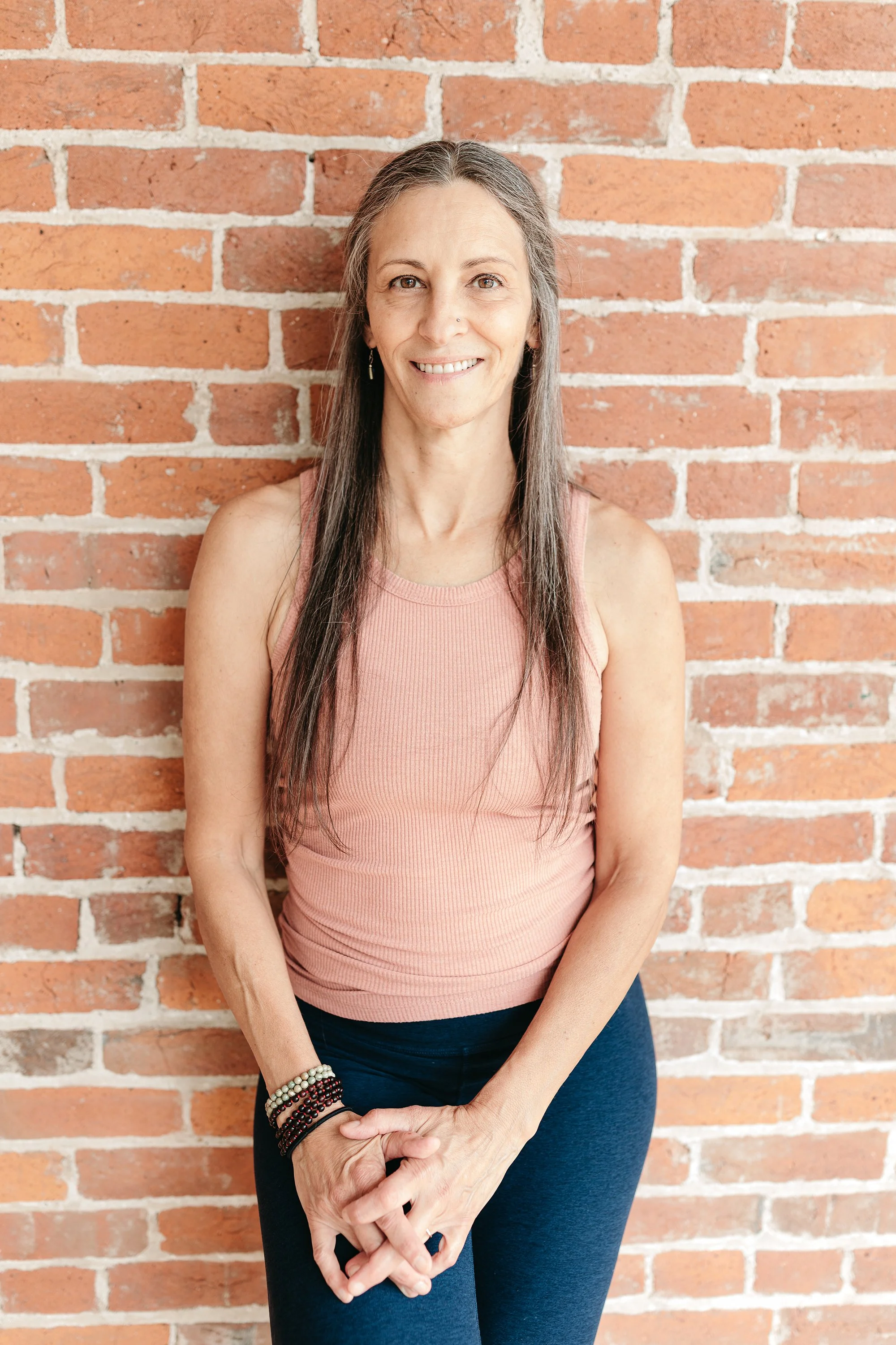 A woman with long dark hair, wearing a blue sleeveless top and gray pants, sitting on a bench in a room with wooden flooring and brick wall background, smiling at the camera.