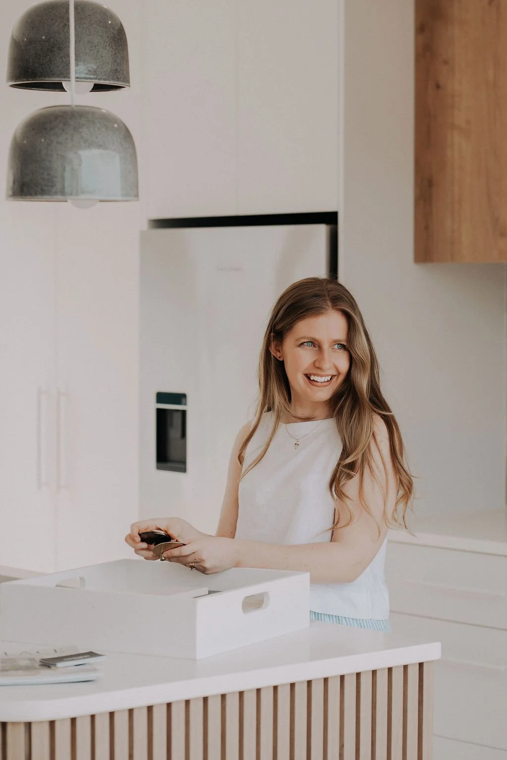 A woman with long light brown hair and blue eyes, wearing a white sleeveless top, smiling and holding a remote control, standing in a modern, minimalistic kitchen with white cabinets, wooden accents, and two hanging pendant lights.