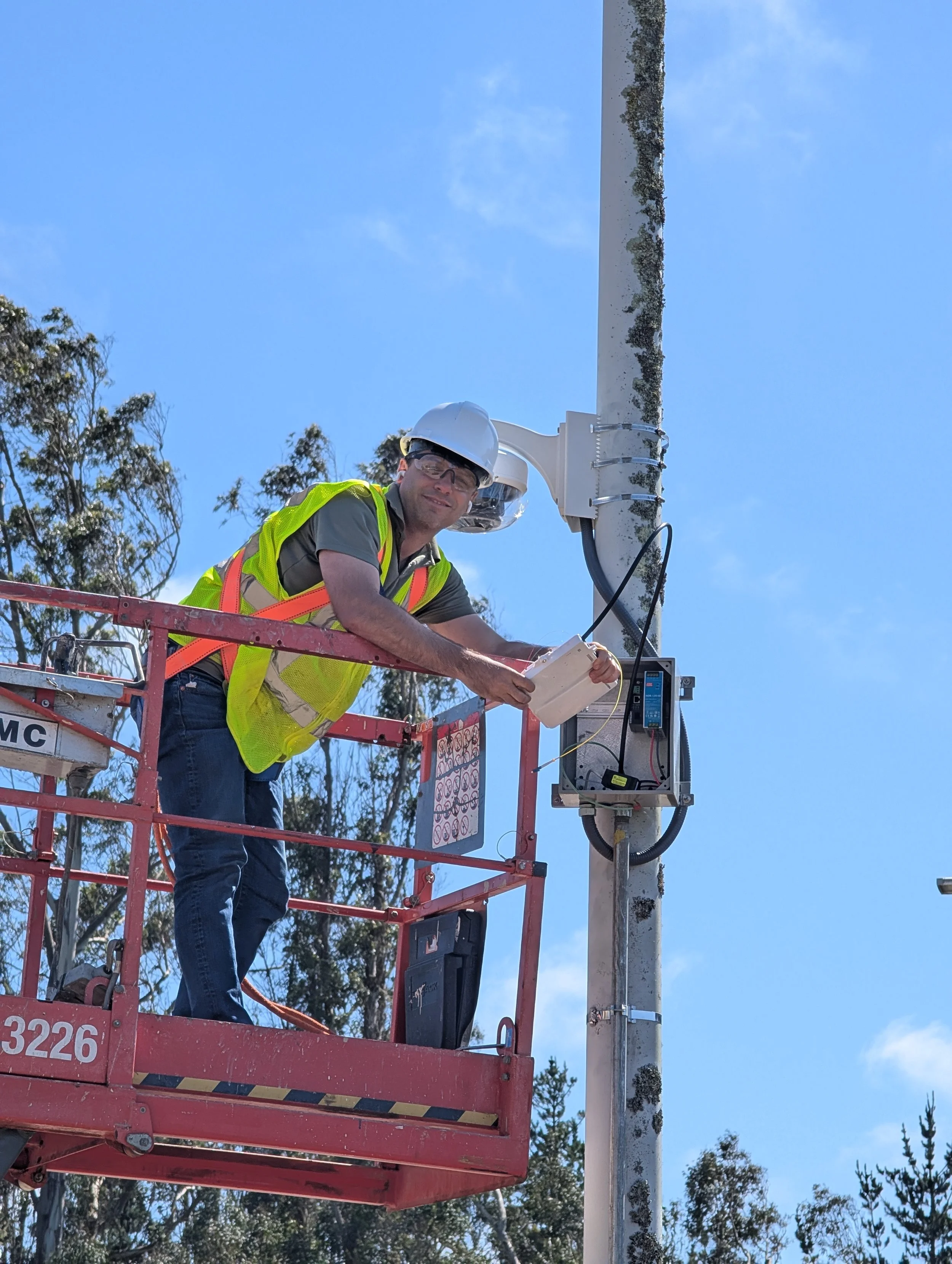 A technician wearing PPE (hard hat, reflective vest, and harness) performs a fiber installation on a scissor lift at a federal facility
