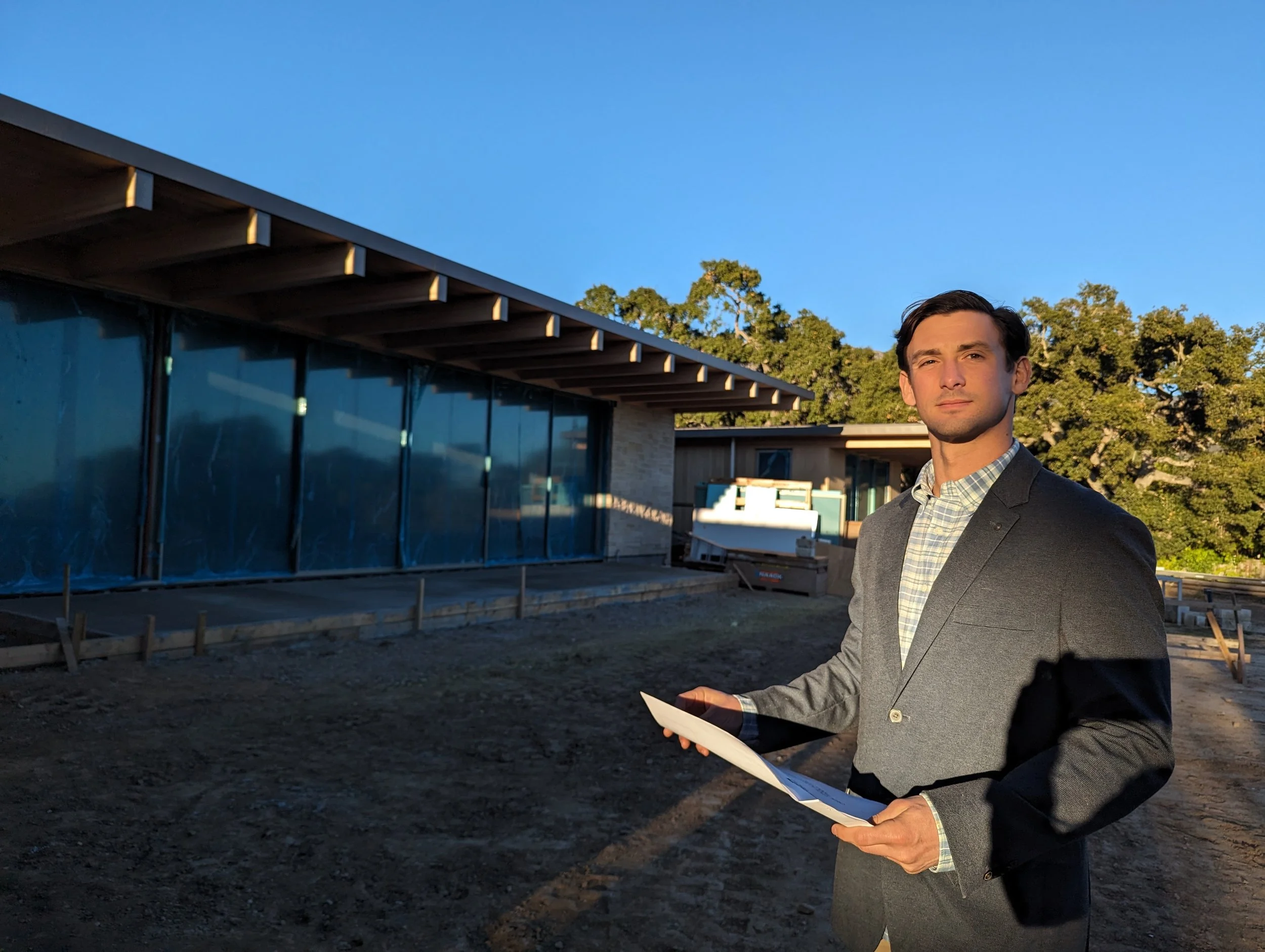 Professional security consultant in a gray blazer holding estate blueprints outside a premium modern residential property during golden hour.