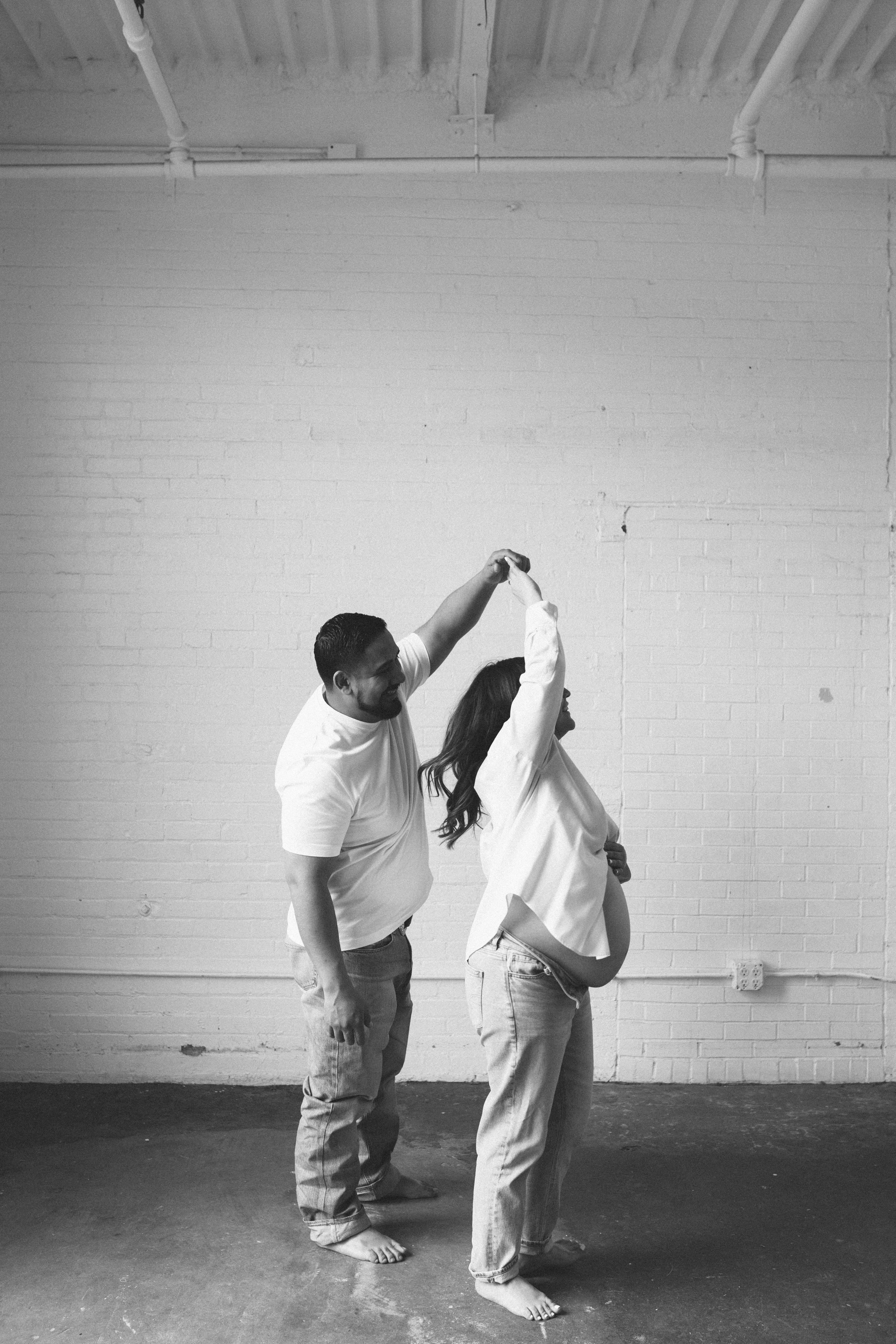 Black and white photo of a man and pregnant woman dancing in a room with bare brick walls.