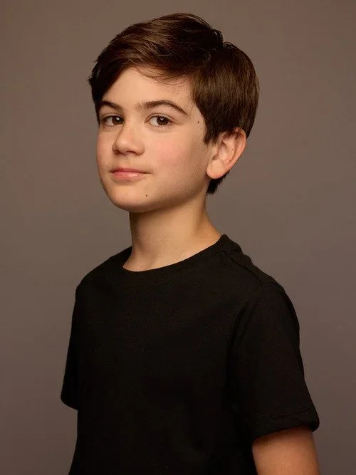 A young boy with short brown hair, wearing a black t-shirt, looking at the camera against a plain gray background.
