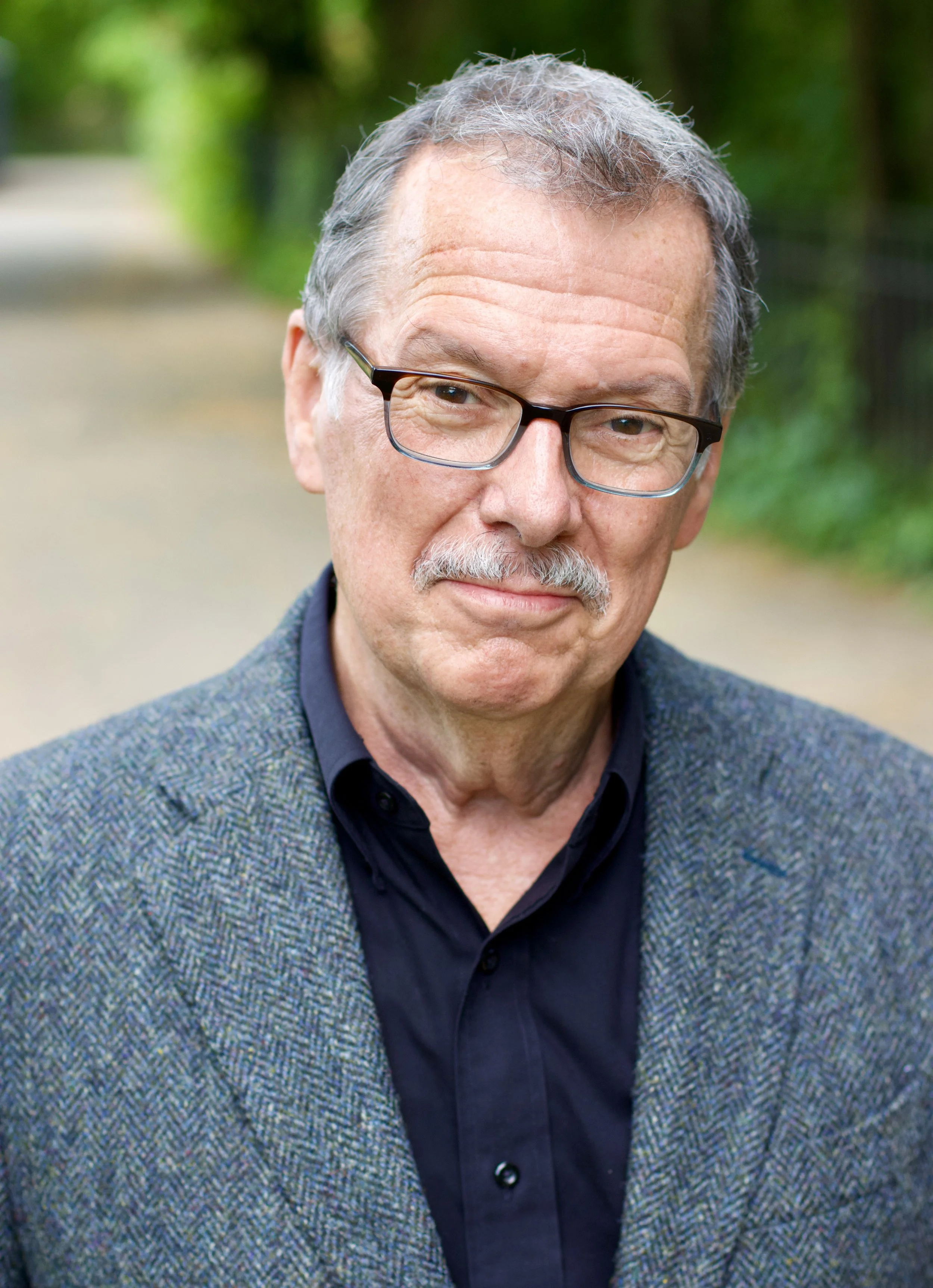 A middle-aged man wearing glasses, a dark shirt, and a gray blazer, standing outdoors with blurred green foliage in the background.