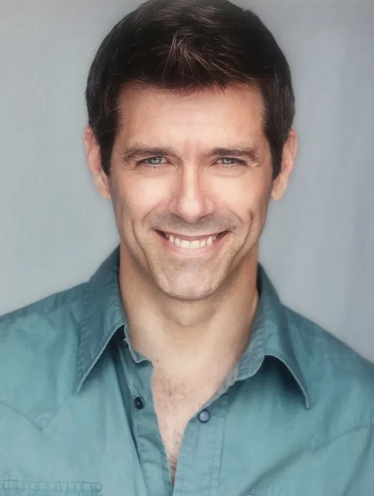 A man with dark hair and light blue eyes, smiling and wearing a teal button-up shirt against a gray background.
