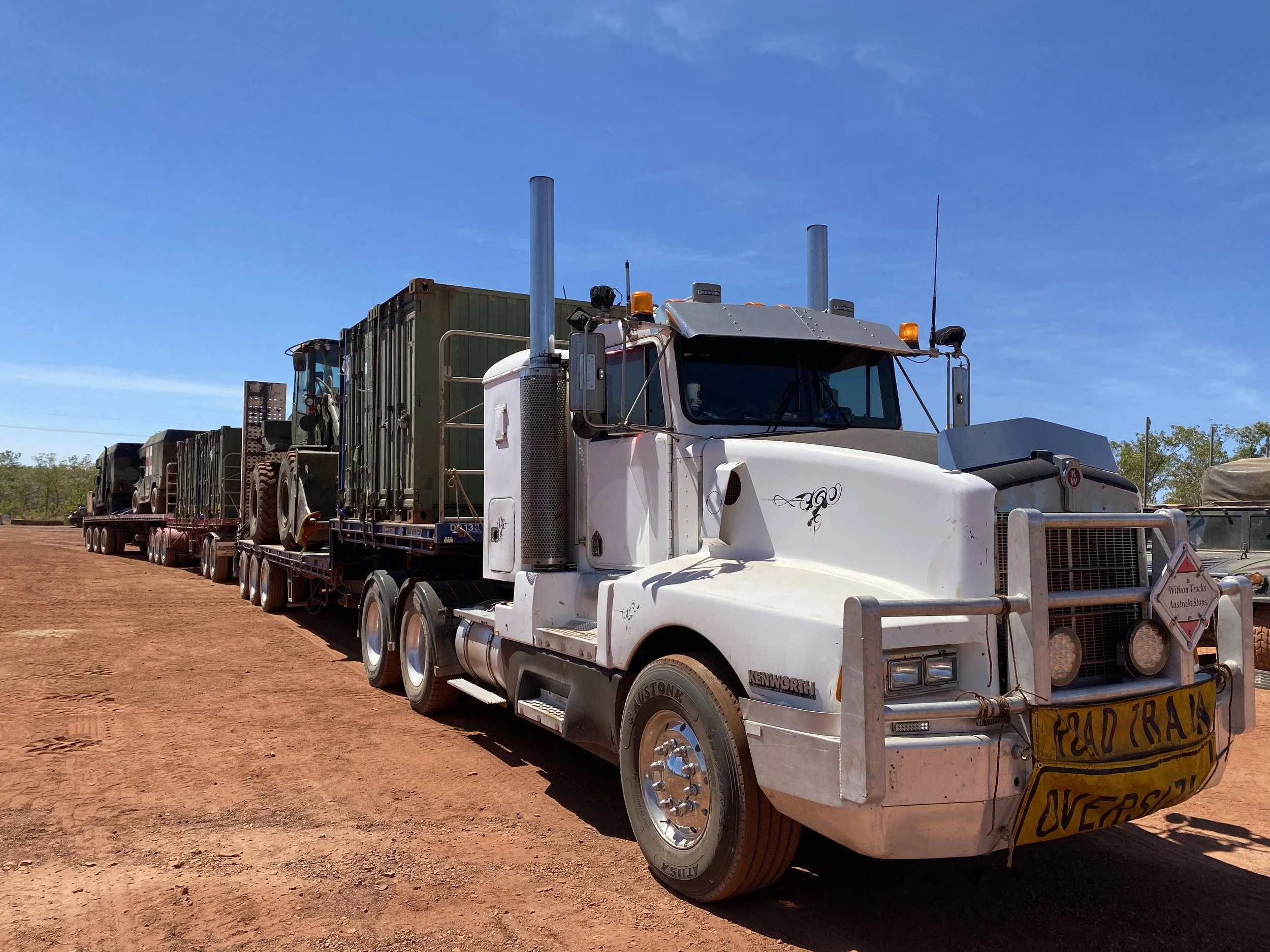 A large white semi-truck with a flatbed trailer carrying military equipment and vehicles on a dirt road under a clear blue sky. The truck is labeled "road train" with a visible oversize load banner.