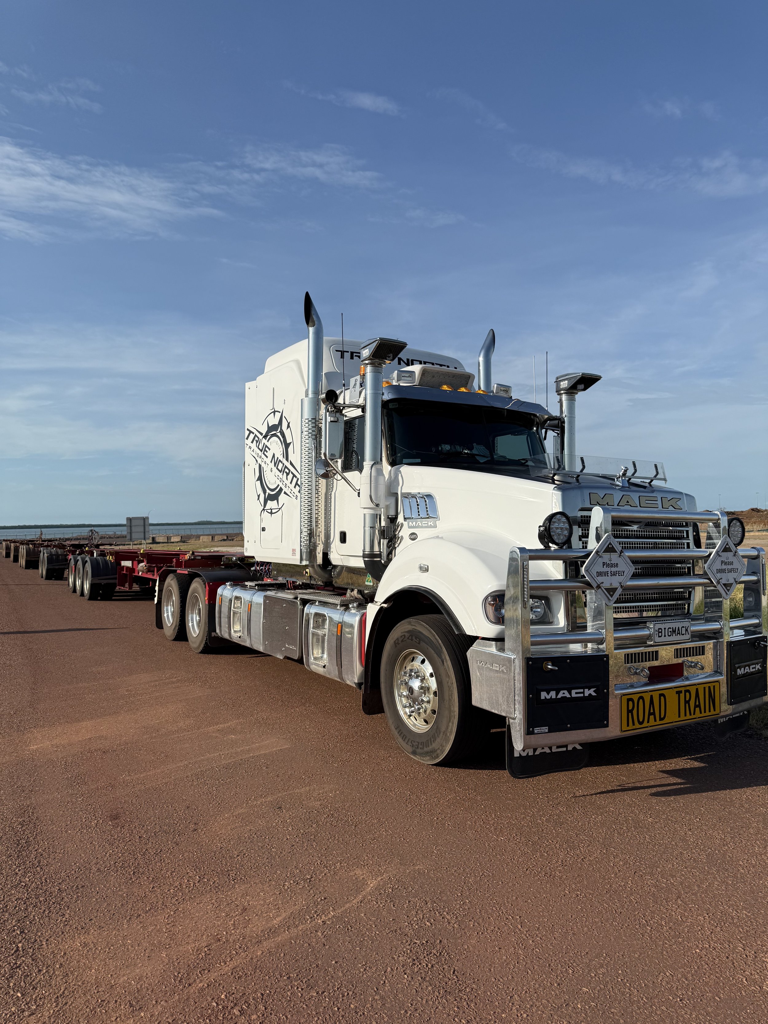 A large white Mack truck labeled "ROAD TRAIN" on a dirt road with clear sky.