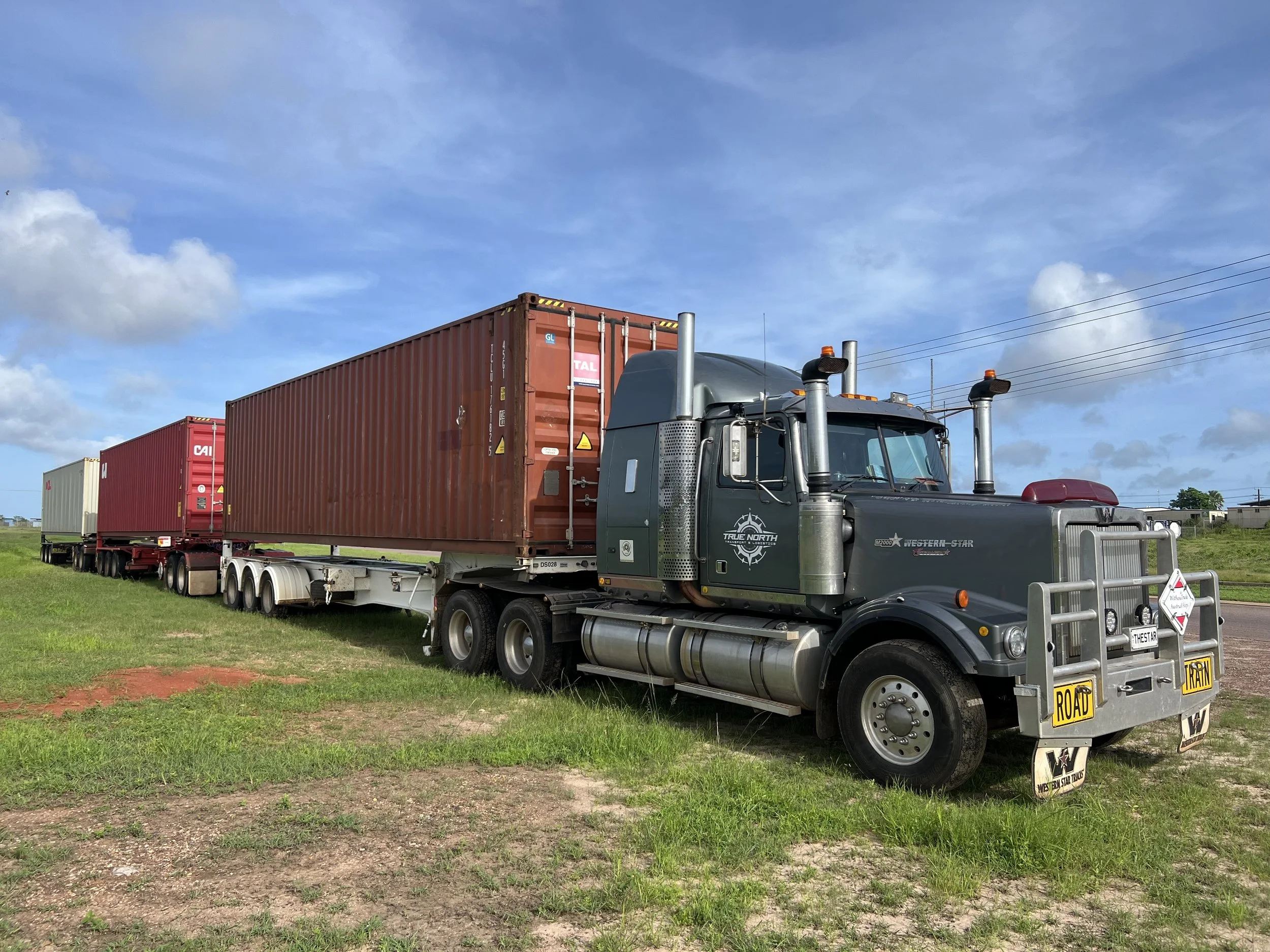 A large semi-truck with a road train configuration, pulling three container trailers on a grassy roadside under a blue sky.