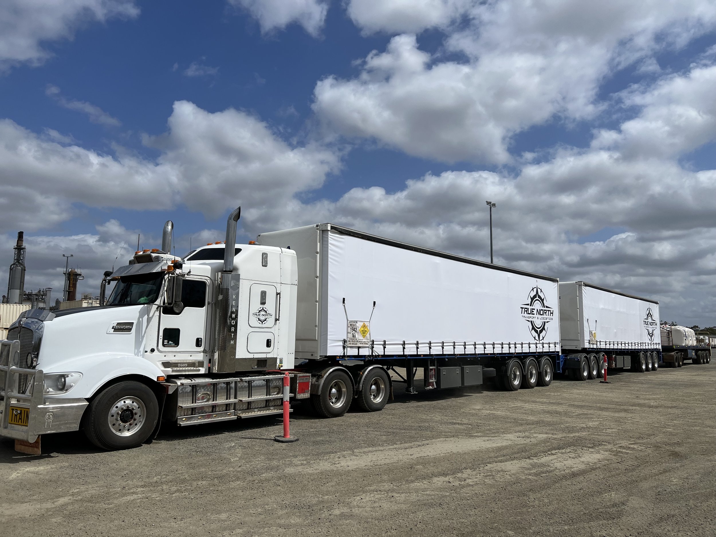 White semi-truck with double trailers parked on a gravel lot under cloudy sky.