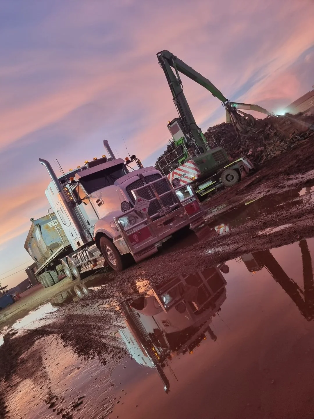 A semi-truck and an excavator in a dirt lot during sunset, with reflections visible in a muddy puddle.