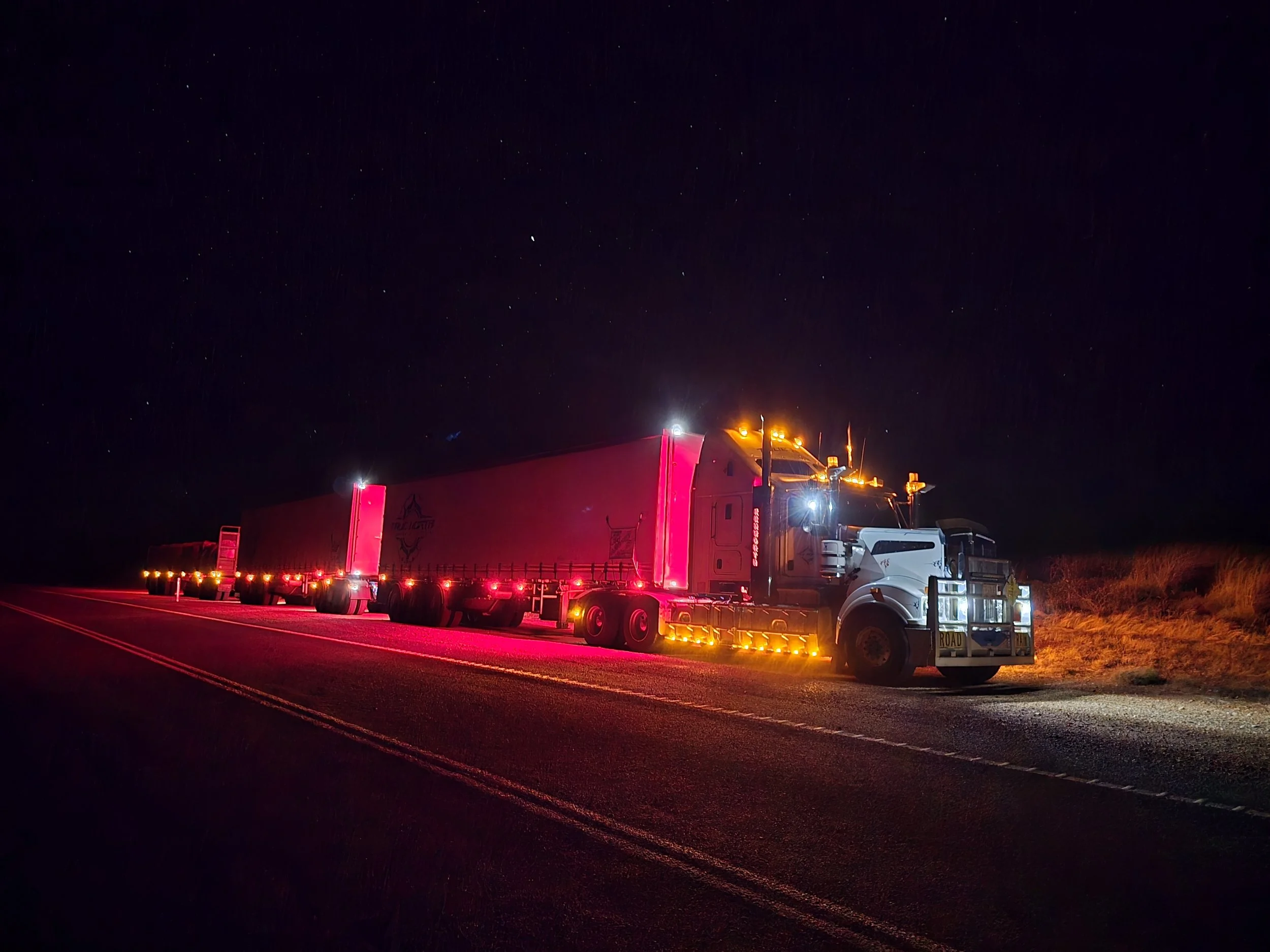 Long-haul road train truck with multiple trailers parked on a highway at night, illuminated by exterior lights.