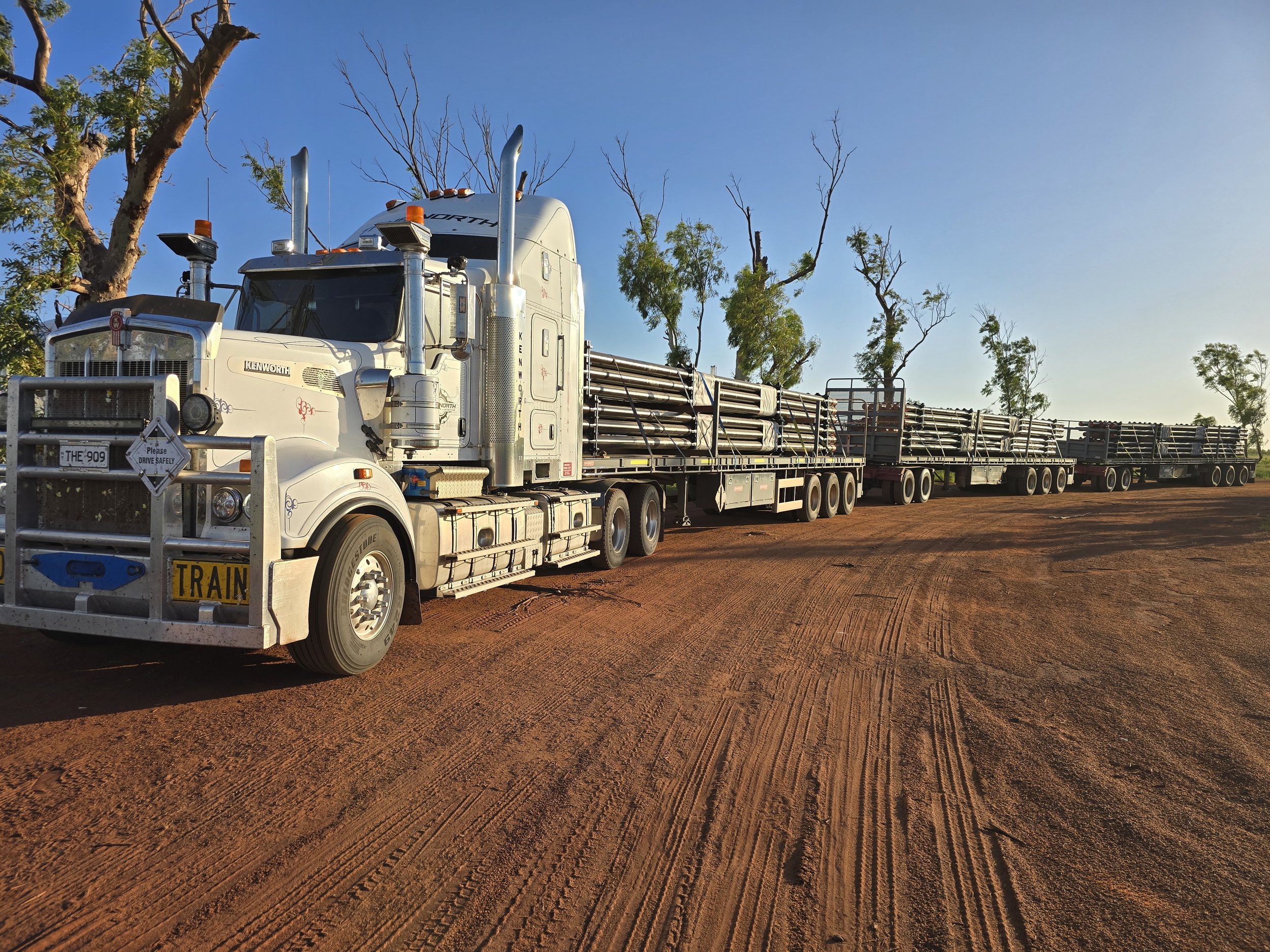 Large road train with multiple trailers on a dirt road with trees in the background under a clear blue sky.