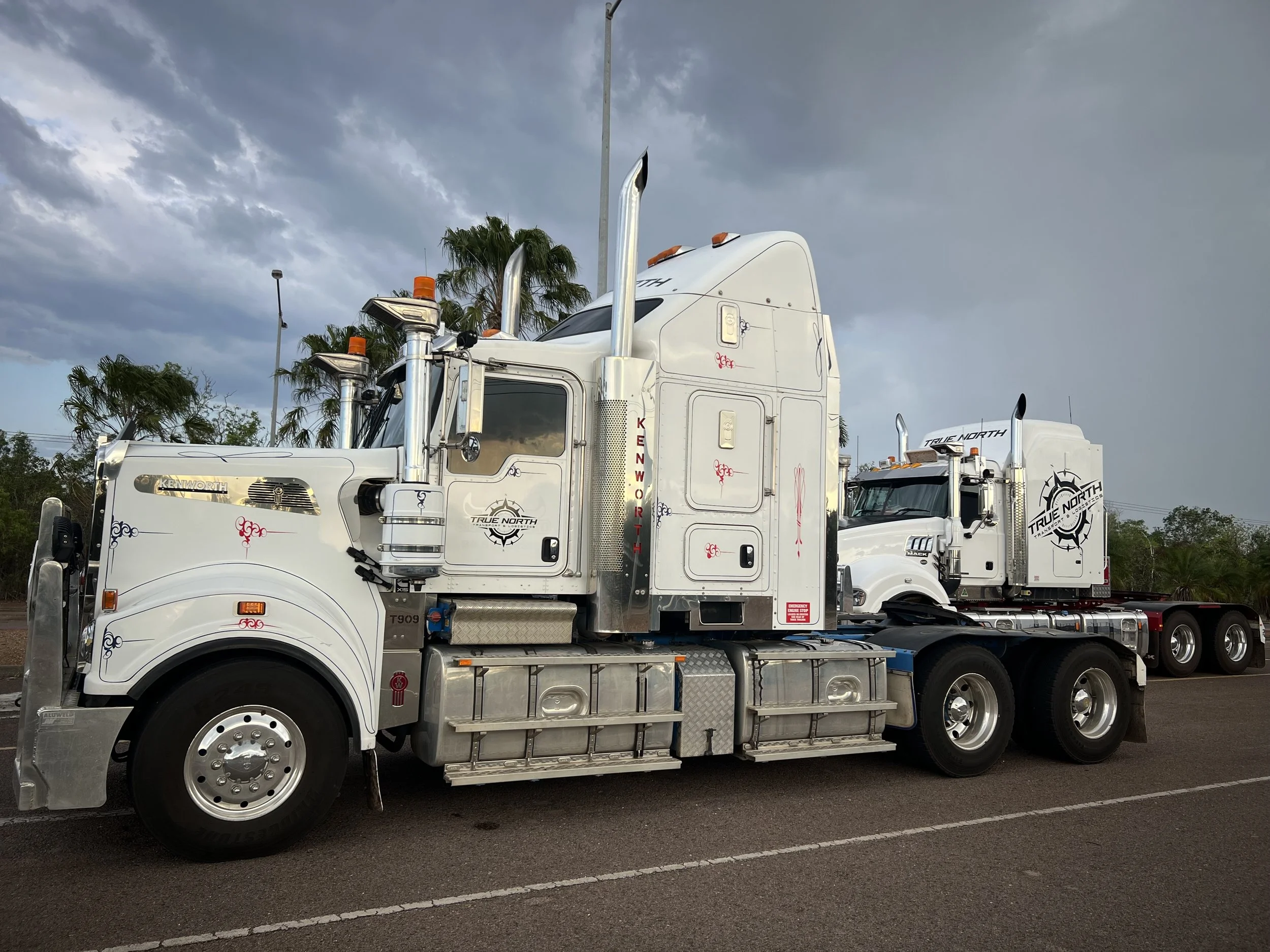 Two white Kenworth semi-trucks parked side by side, with "True North" branding, under a cloudy sky with palm trees in the background.