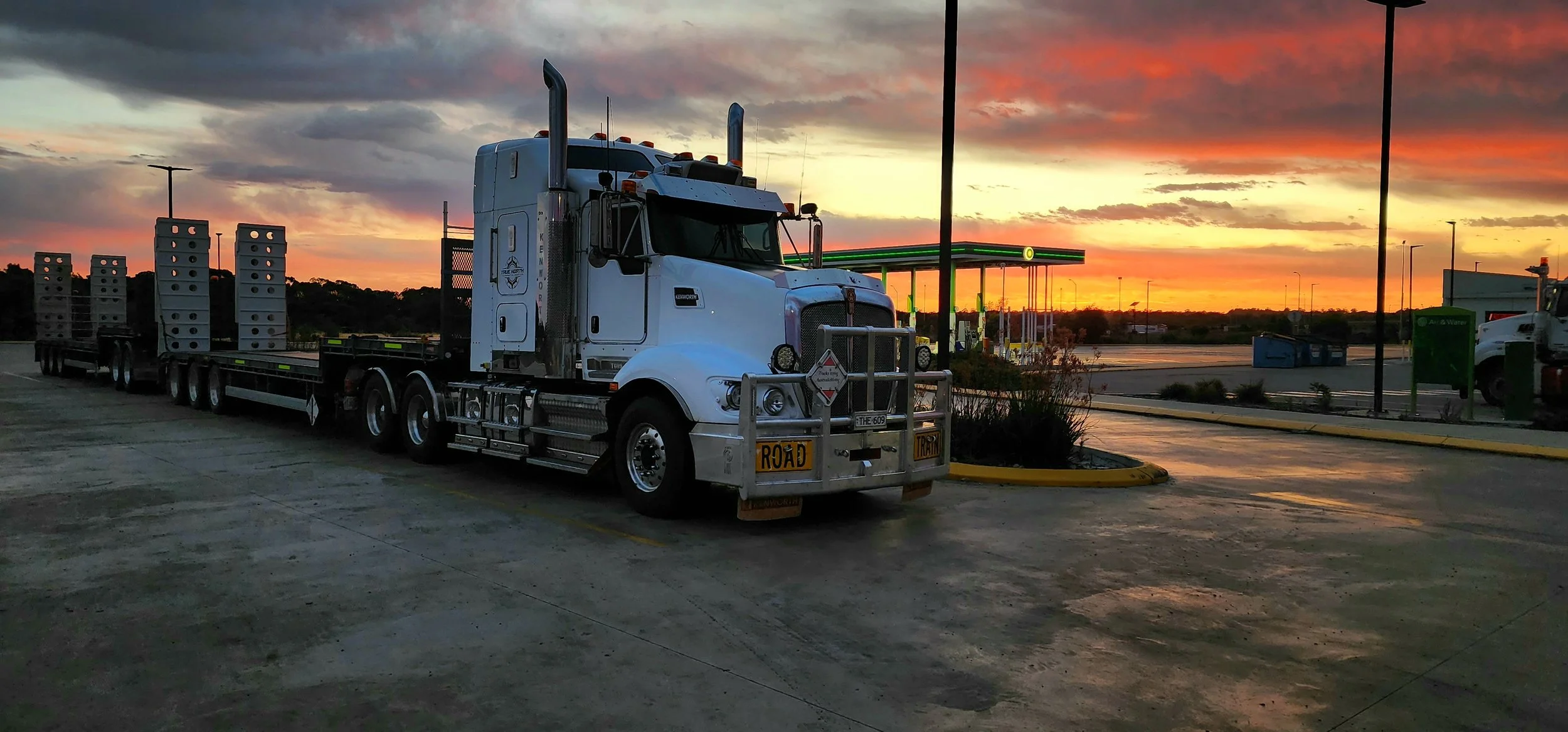 White semi-truck with flatbed trailer parked at a gas station during a vibrant sunset.
