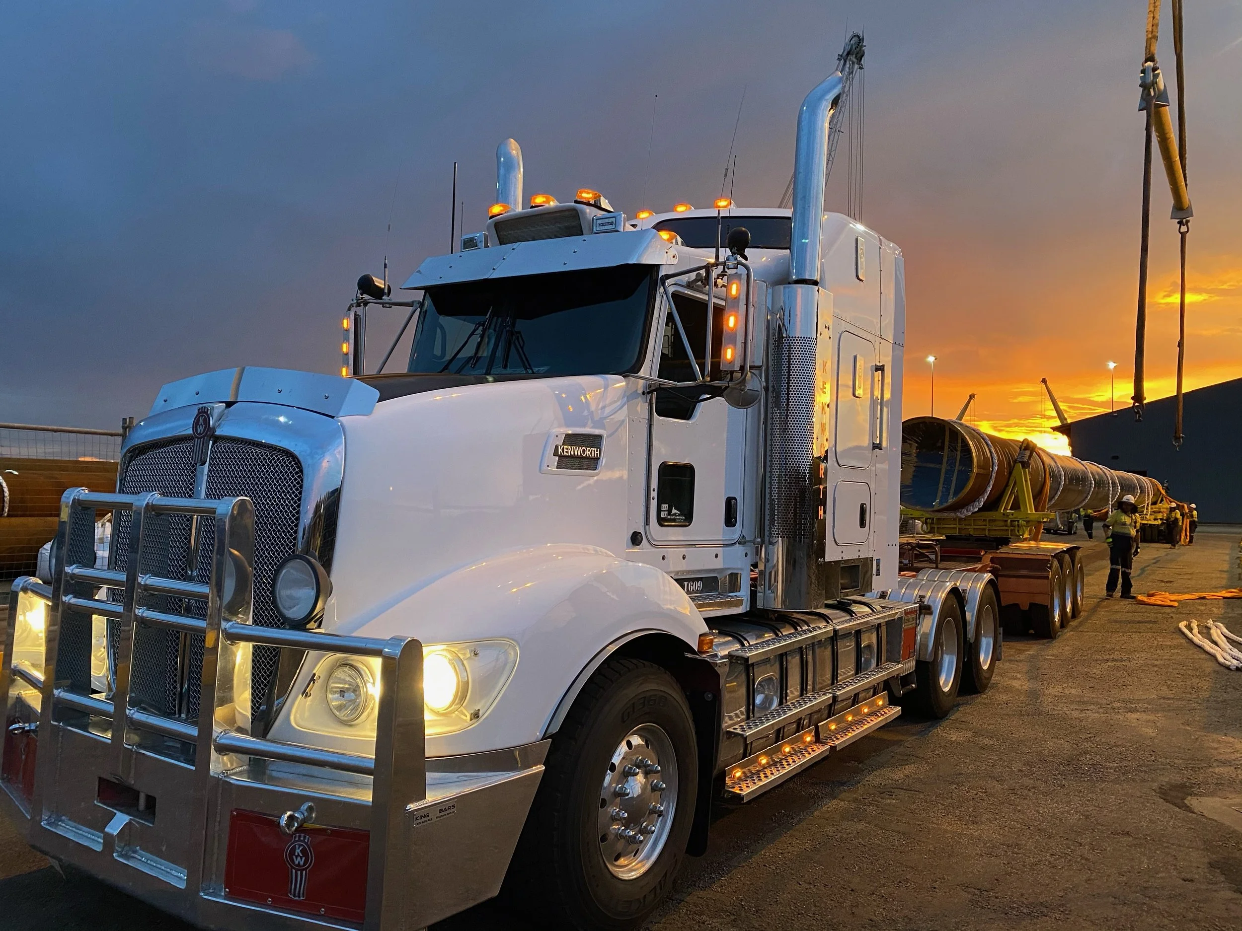 A white Kenworth semi-truck parked with a large trailer carrying long cylindrical pipes at an industrial site during sunset. Workers are visible in the background.