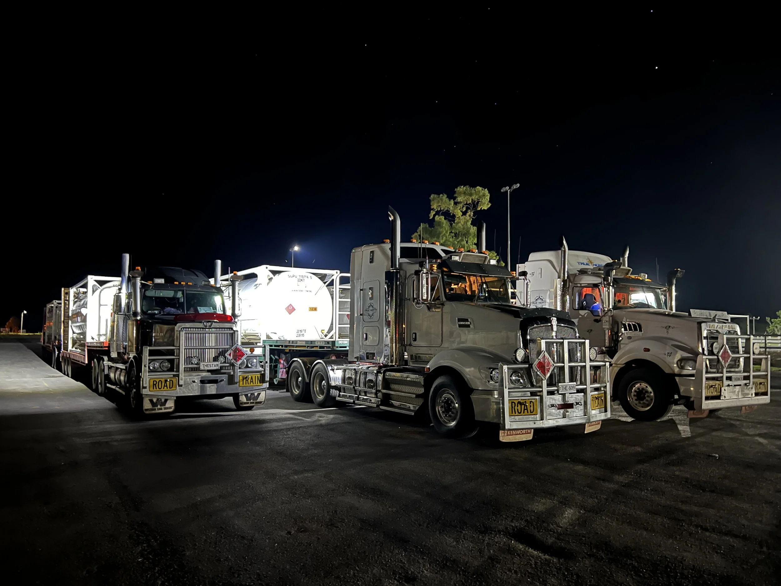 Three large road train trucks parked at night, each with a prominent "ROAD TRAIN" sign on the front, under a dark sky.
