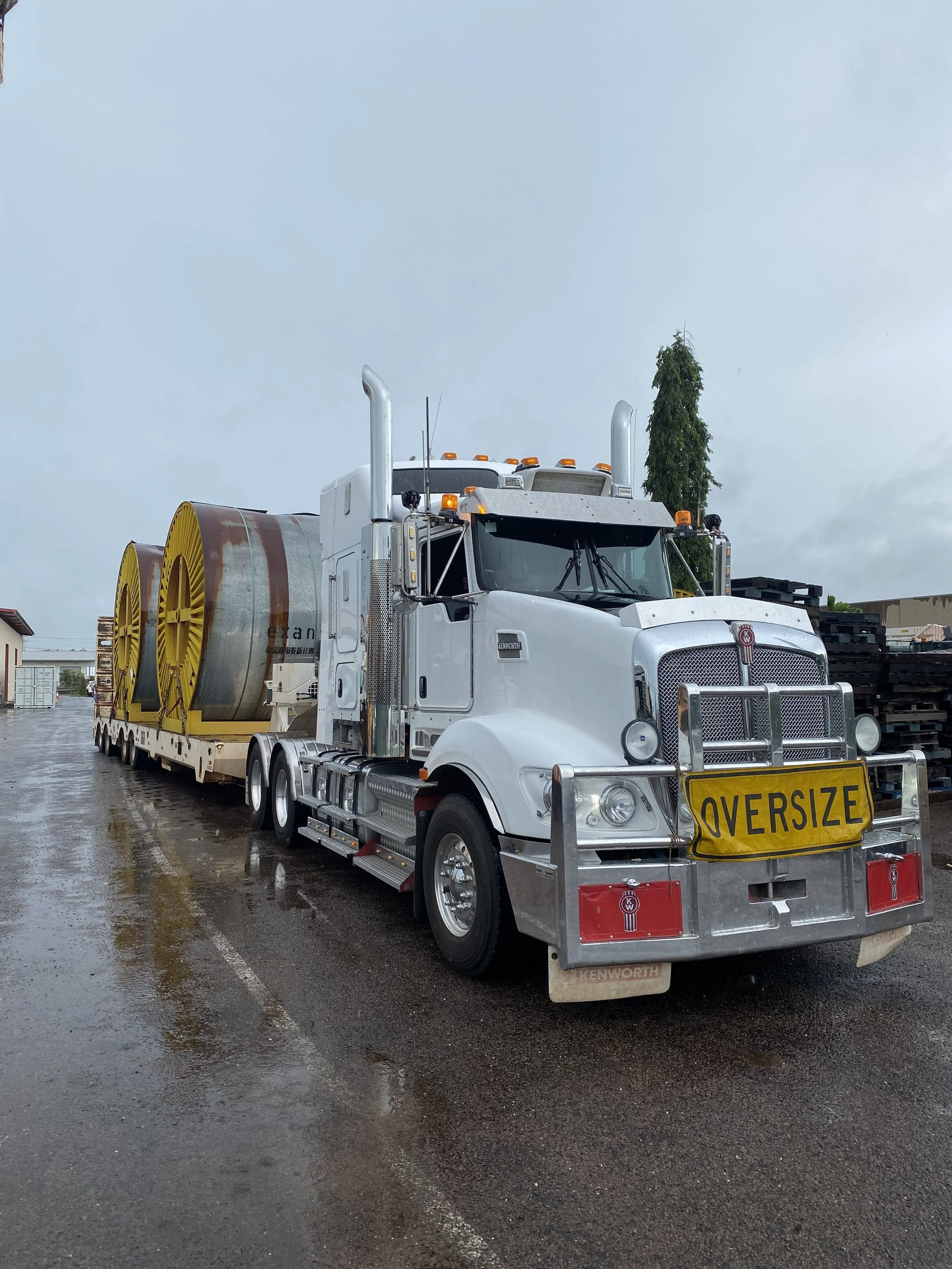 Large white semi-truck with "OVERSIZE" sign, pulling a flatbed trailer carrying large industrial equipment spools, on a wet roadway.