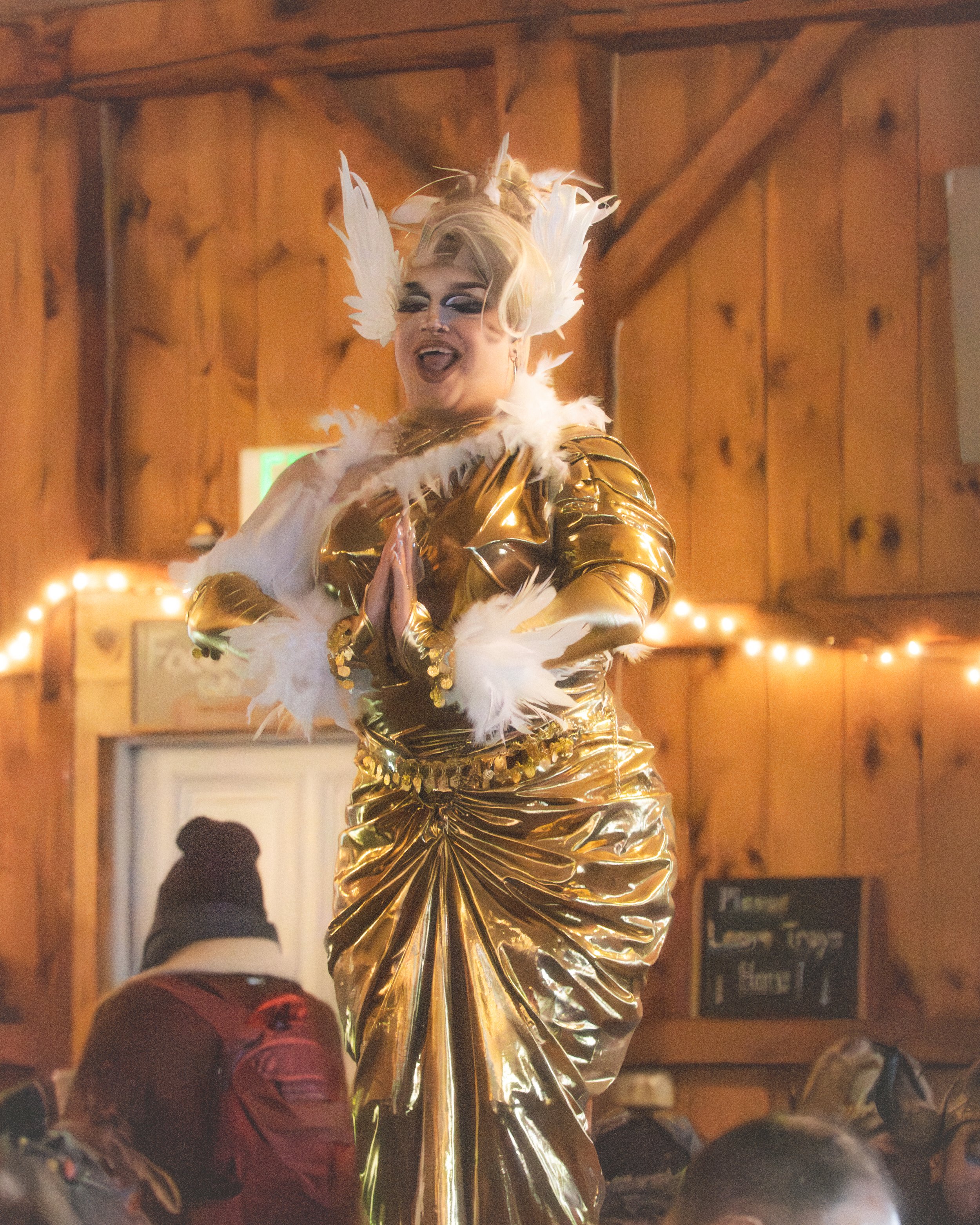 A drag performer dressed in a gold, metallic gown with white feather accents, wearing large feathered headpiece, performing in a warmly lit wood-paneled room.