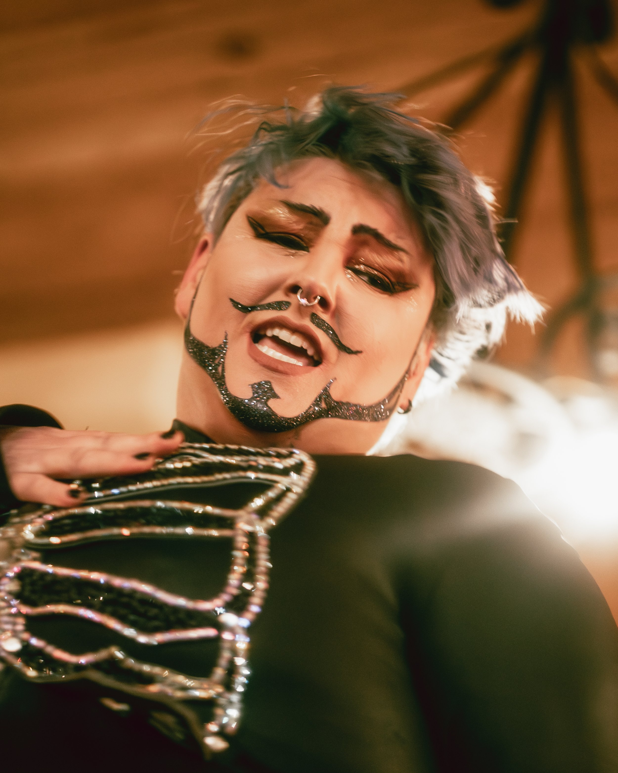 Person with makeup and glitter facial design, short tousled hair, wearing multiple chains, in a pose with hand on chest, in a warm indoor setting.