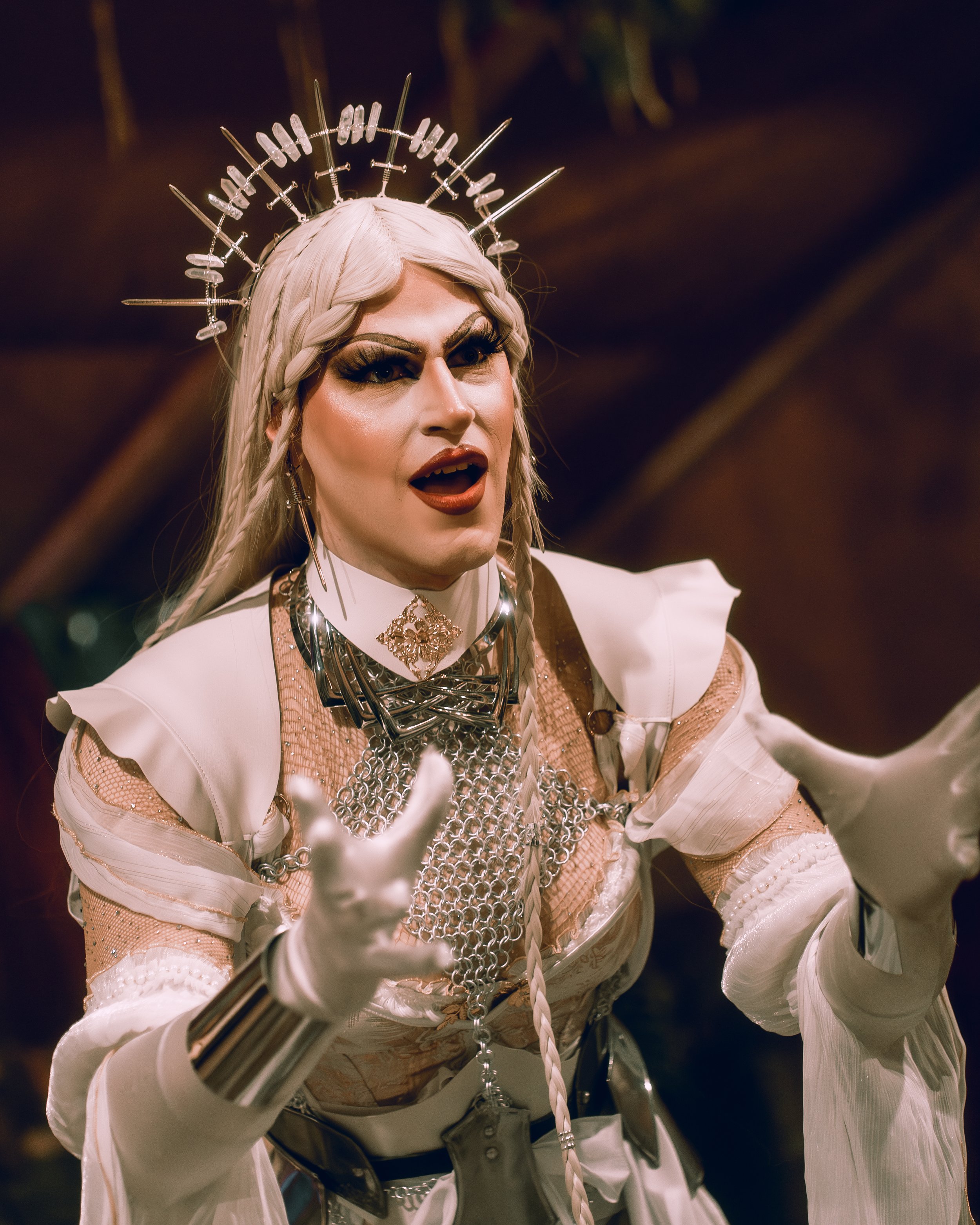 A person dressed in elaborate, futuristic costume with armored white gloves, intricate jewelry, and braided hair. They are wearing a silver halo and appear to be speaking or singing passionately.