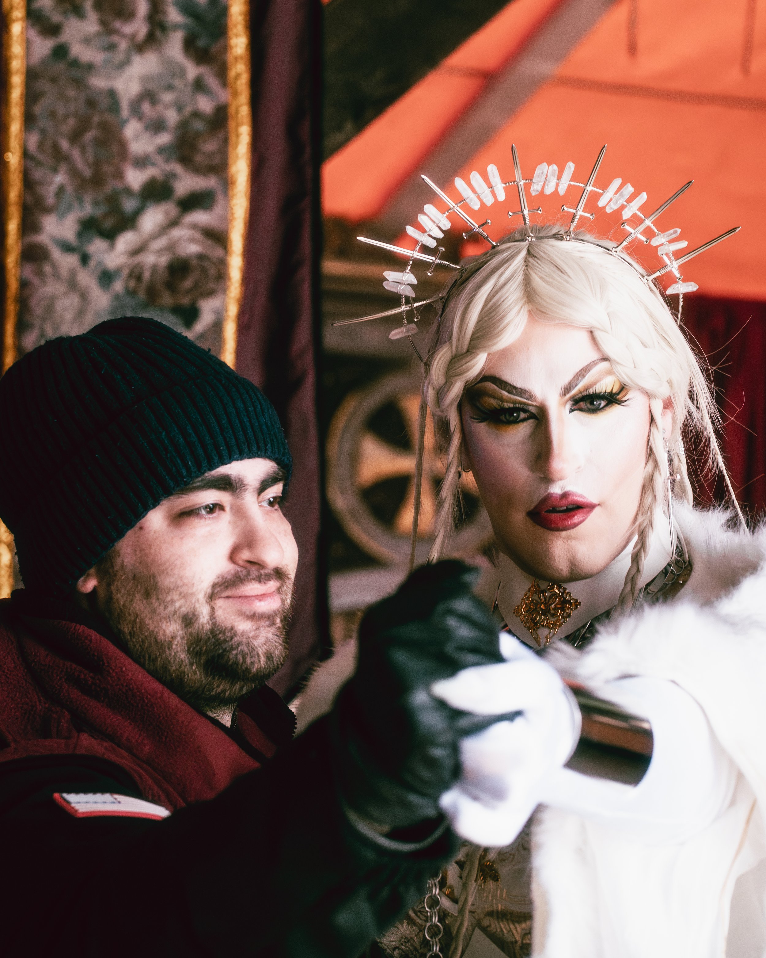 A man and a drag queen pose for a photo inside a decorated room. The man wears a black beanie and burgundy hoodie, while the drag queen has platinum blonde hair, dramatic makeup, and a halo headpiece made of metal sticks and stones, dressed in white 