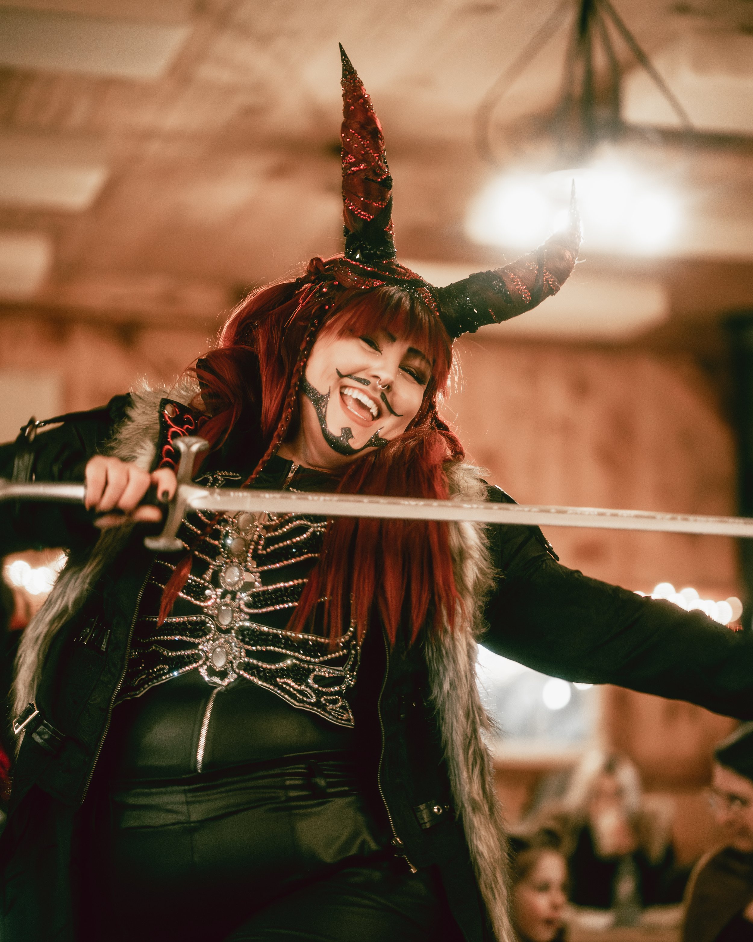 A woman with red hair wearing a black leather outfit and a fur vest, smiling and holding a sword, in a festive or costume setting with horned headgear and face paint.