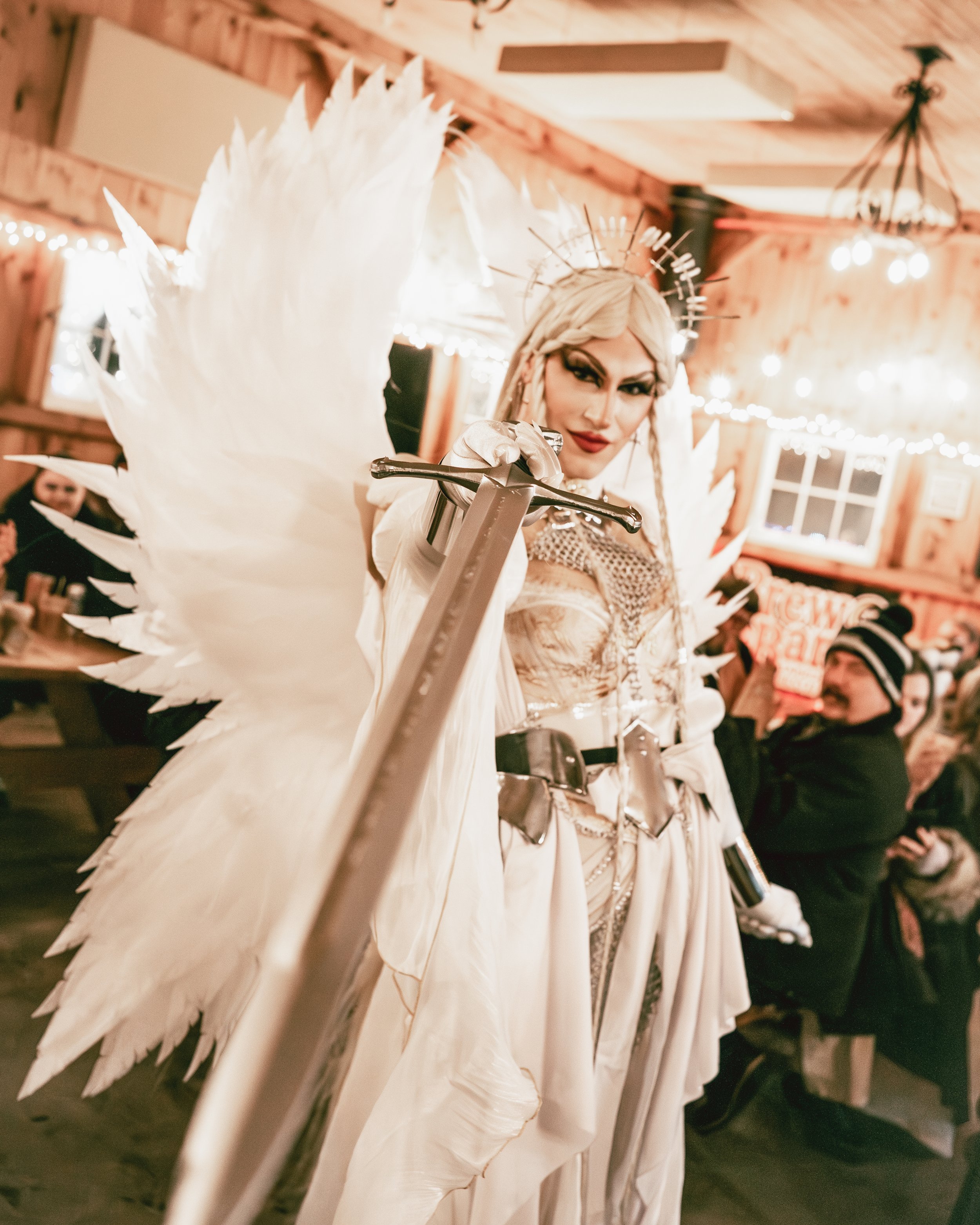 Person dressed as an angel with large white wings, crown, and costume, holding a sword, at a festive indoor event with wooden walls, fairy lights, and seated guests.