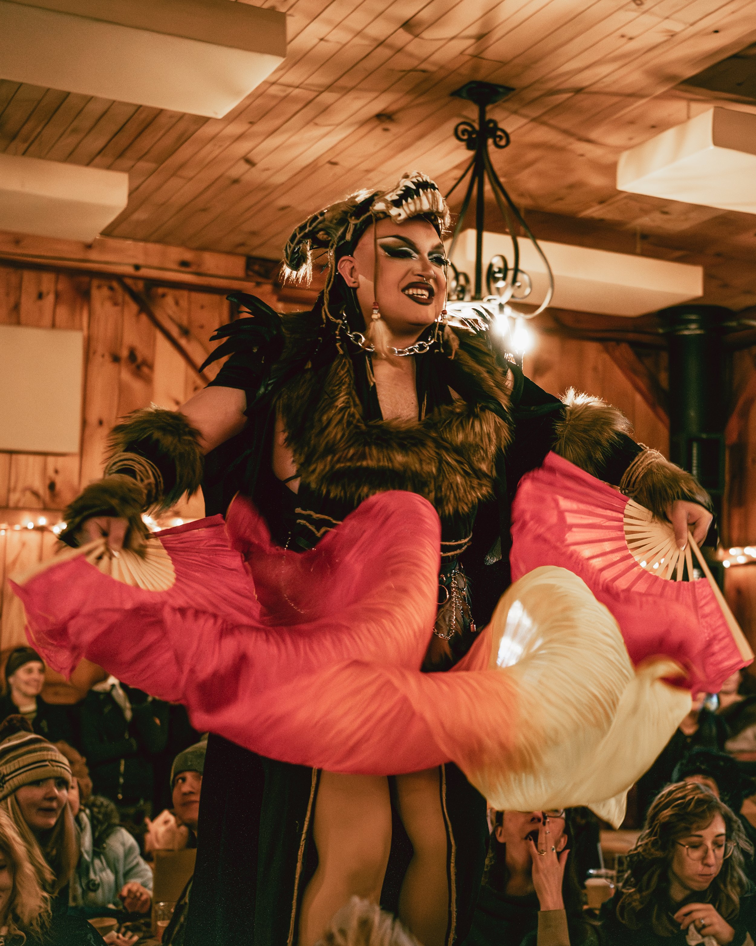 A performer with elaborate makeup, wearing a feathered costume, holding pink and cream fans, on a stage in a wooden hall.