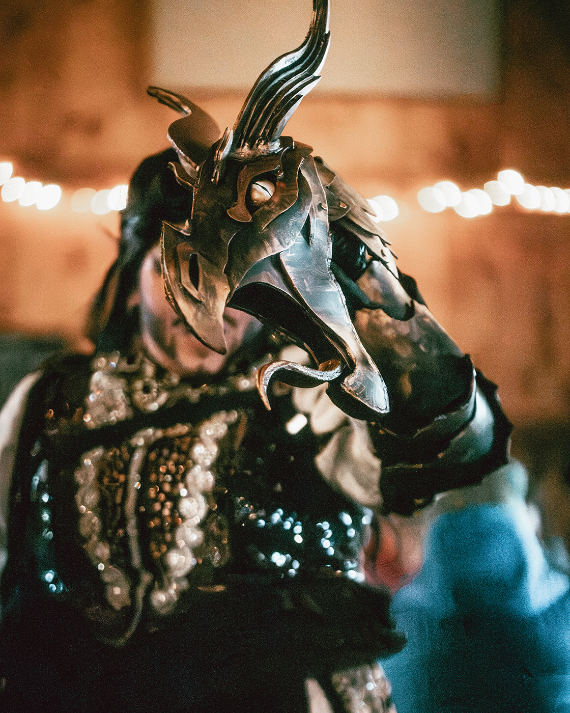 A person wearing an elaborate metallic mask and costume, with a dark background and string lights.