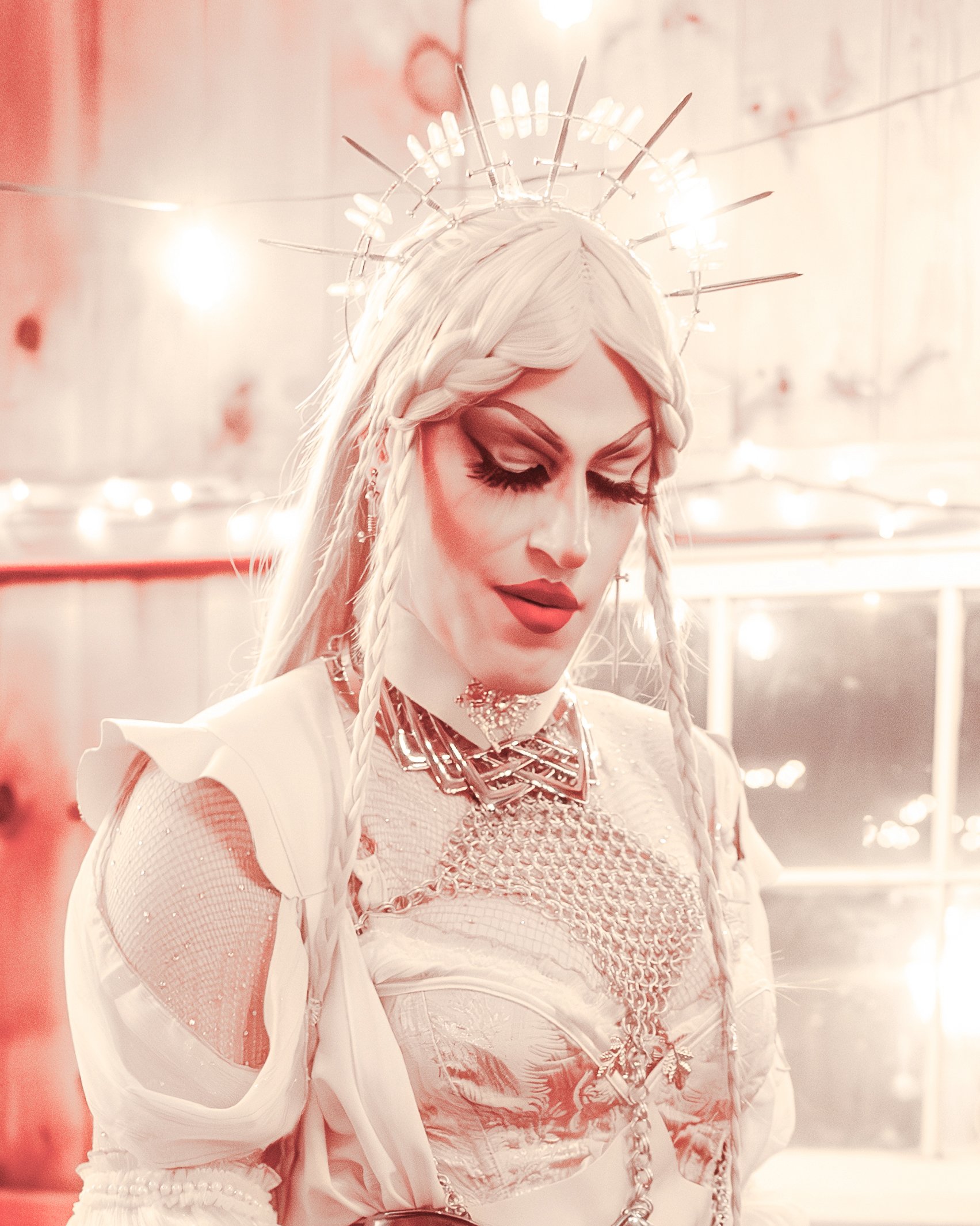 A person dressed in elaborate, futuristic fashion with platinum blonde braided hair, dramatic makeup, and silver jewelry, standing indoors with warm lighting.