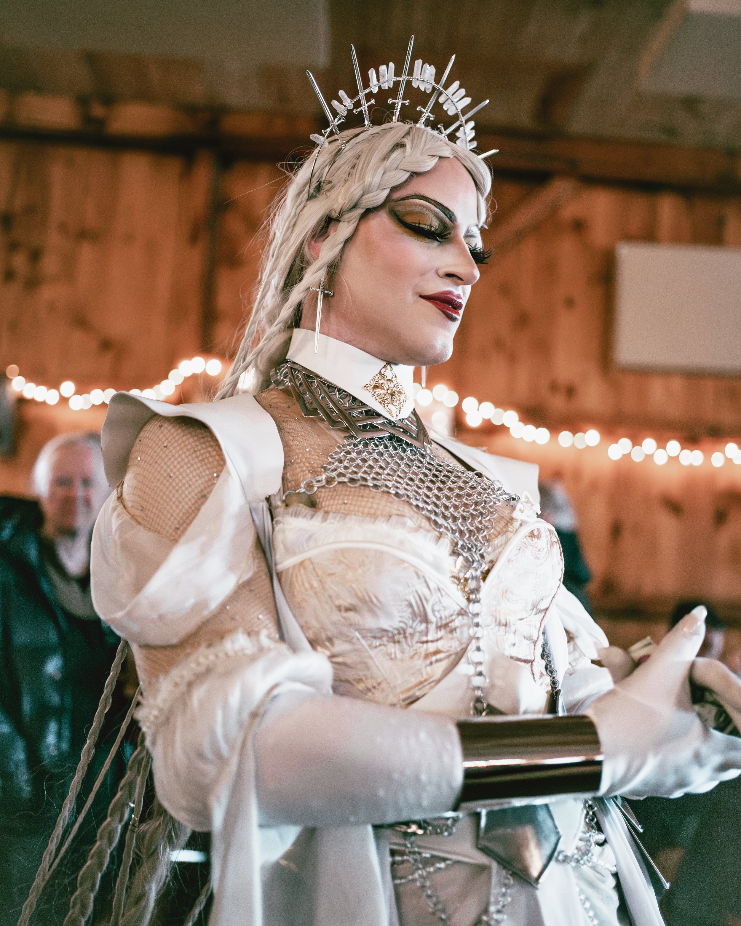 A woman dressed in elaborate, futuristic attire with metallic accessories, jewelry, and makeup, wearing a silver crown with spikes, in a warmly lit wooden indoor setting.