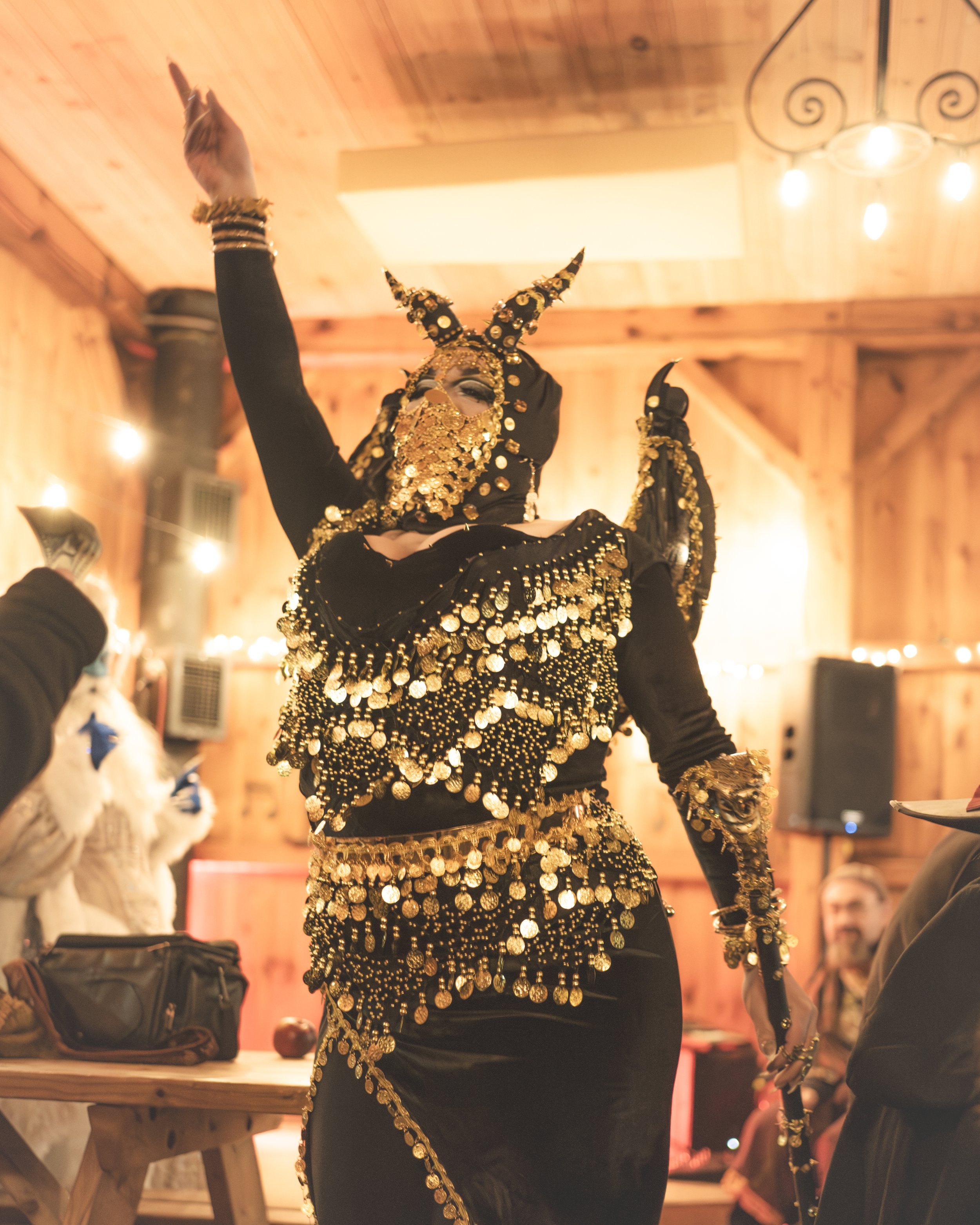 Person dressed in a black costume with gold coins and ornaments, wearing a mask with horns, performing at a gathering in a wooden indoor setting.