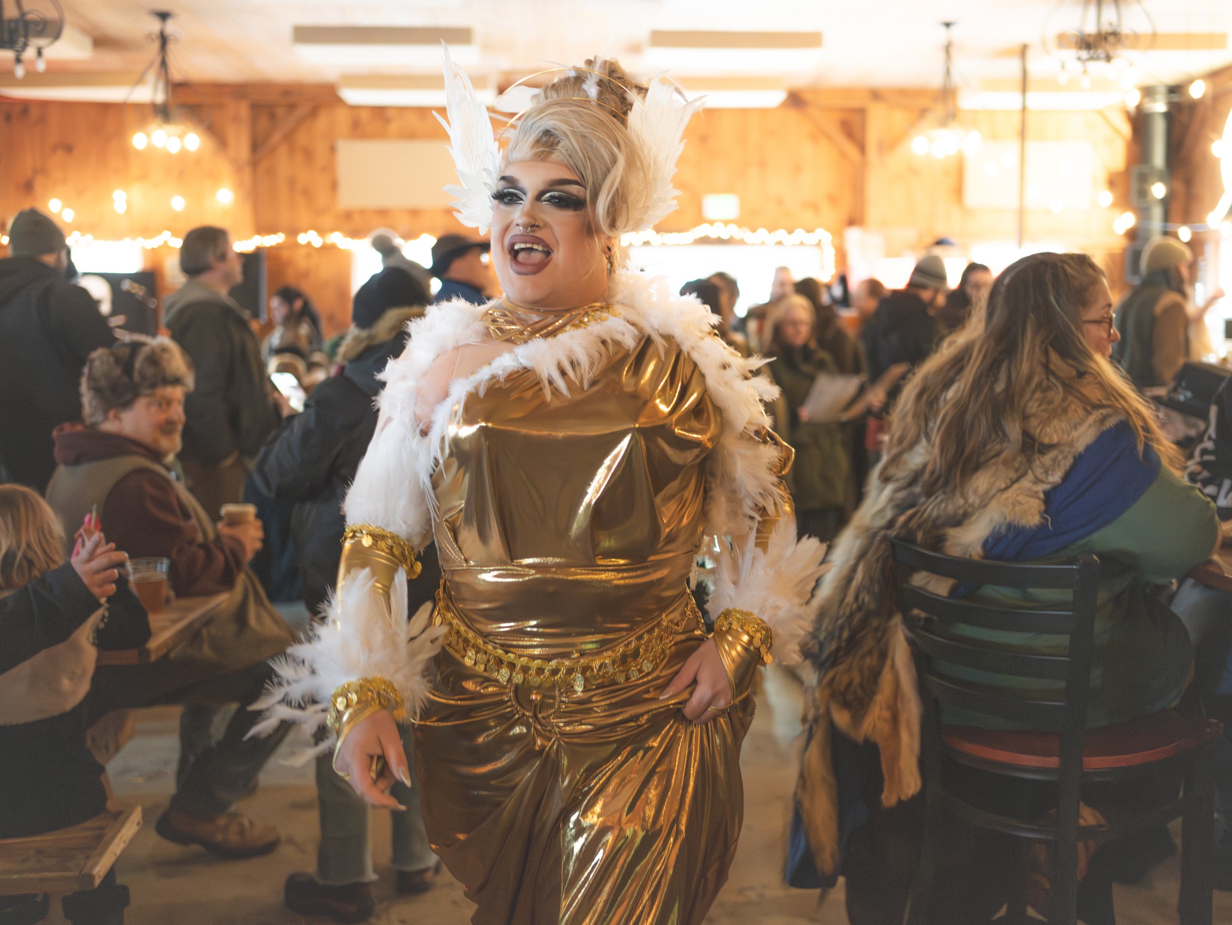 A drag queen with platinum blonde hair, elaborate makeup, white feather headpiece, and a gold metallic dress with white feather trim, standing in a lively indoor event space with people in the background.