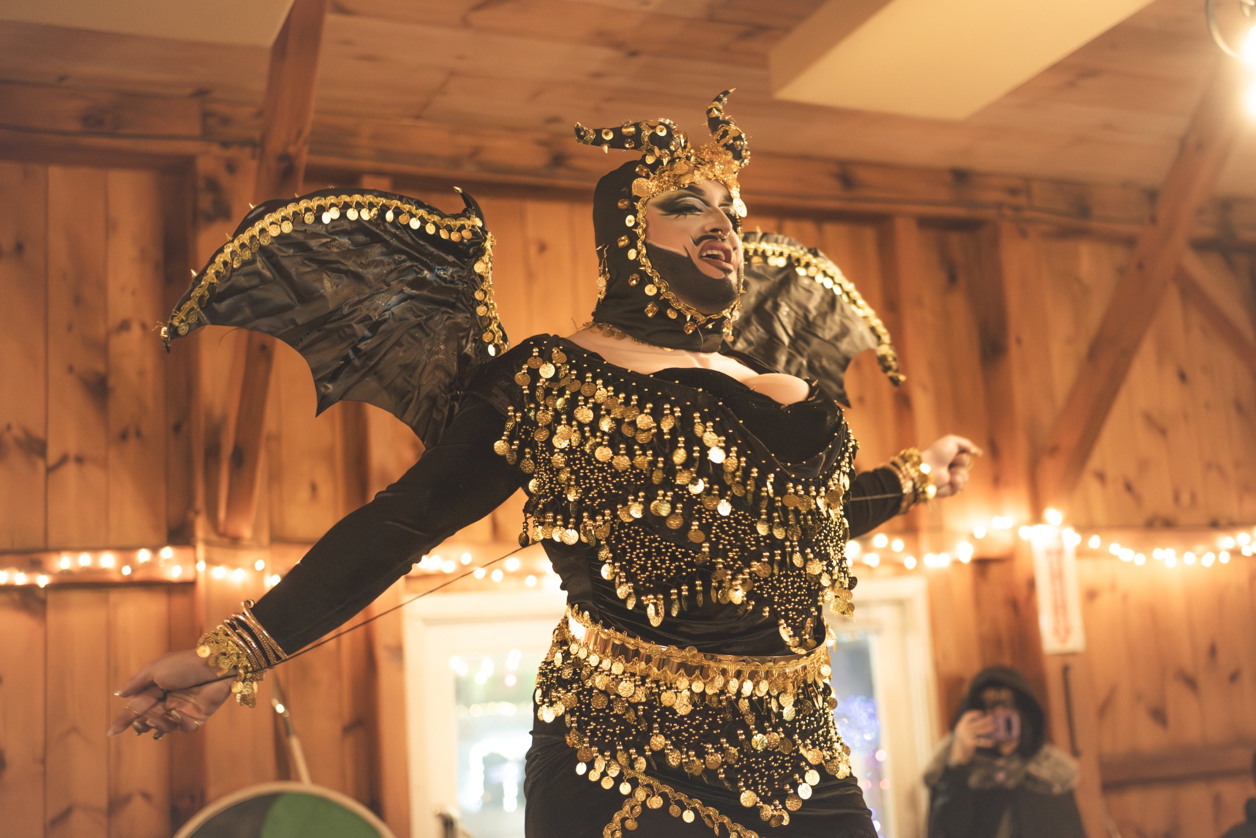 Performer dressed as a dragon with black and gold costume, wings, and horned headpiece, singing or speaking passionately in a wooden indoor setting with string lights, with a person taking photos in the background.