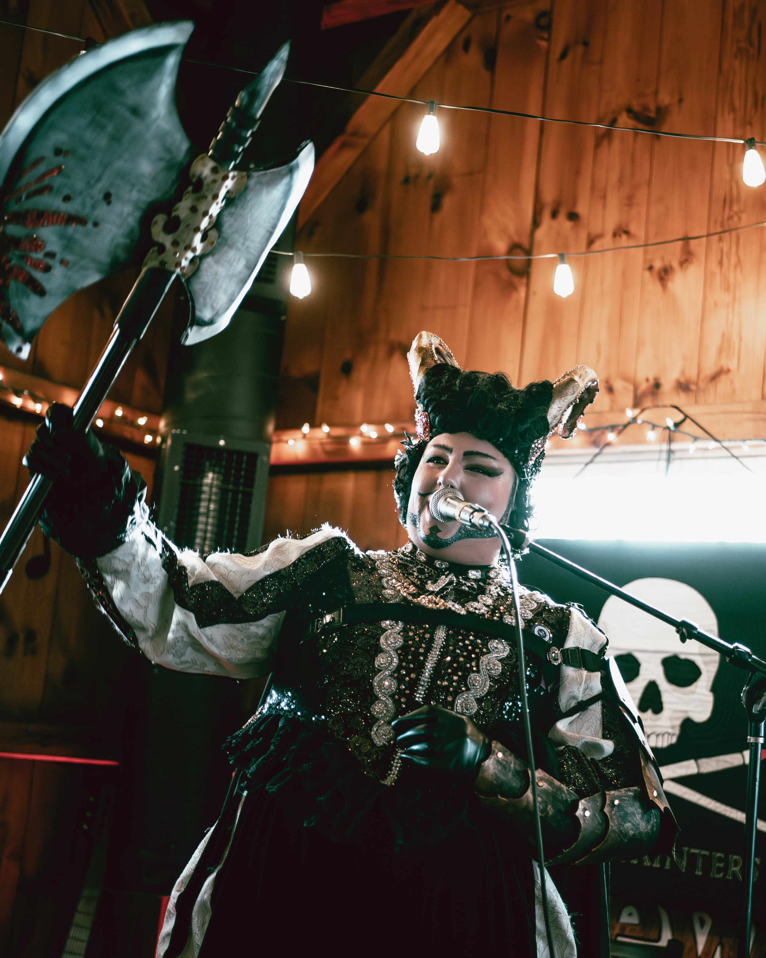A woman dressed as a mythical sea creature or witch, with a headdress featuring seashells and animal ears, singing into a microphone, holding a large prop resembling a seahorse or mythical creature's horn, with a backdrop featuring a skull flag and w
